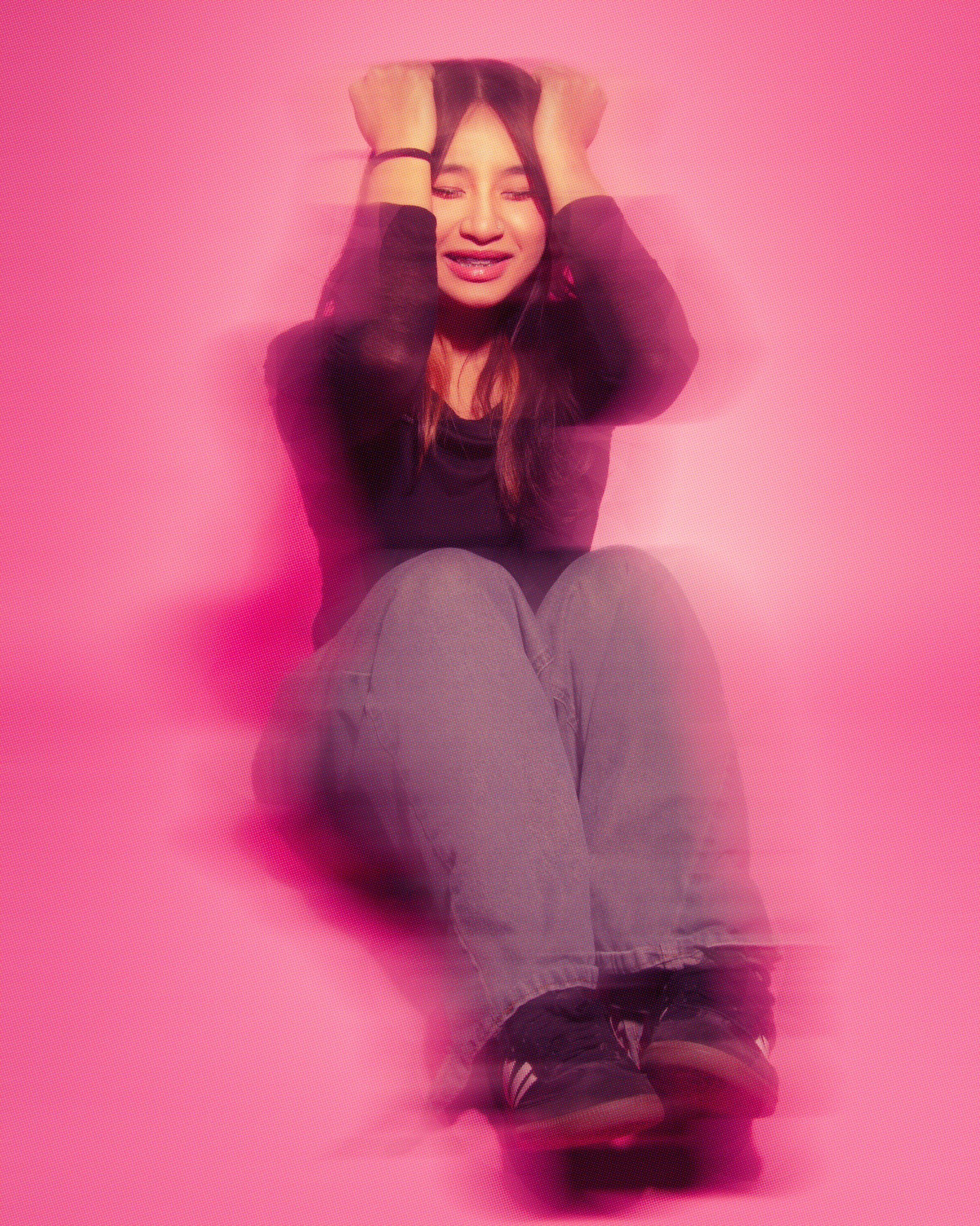 A person sitting against a pink wall, holding their head with both hands, smiling slightly, wearing black long-sleeve top, beige pants, and black sneakers.