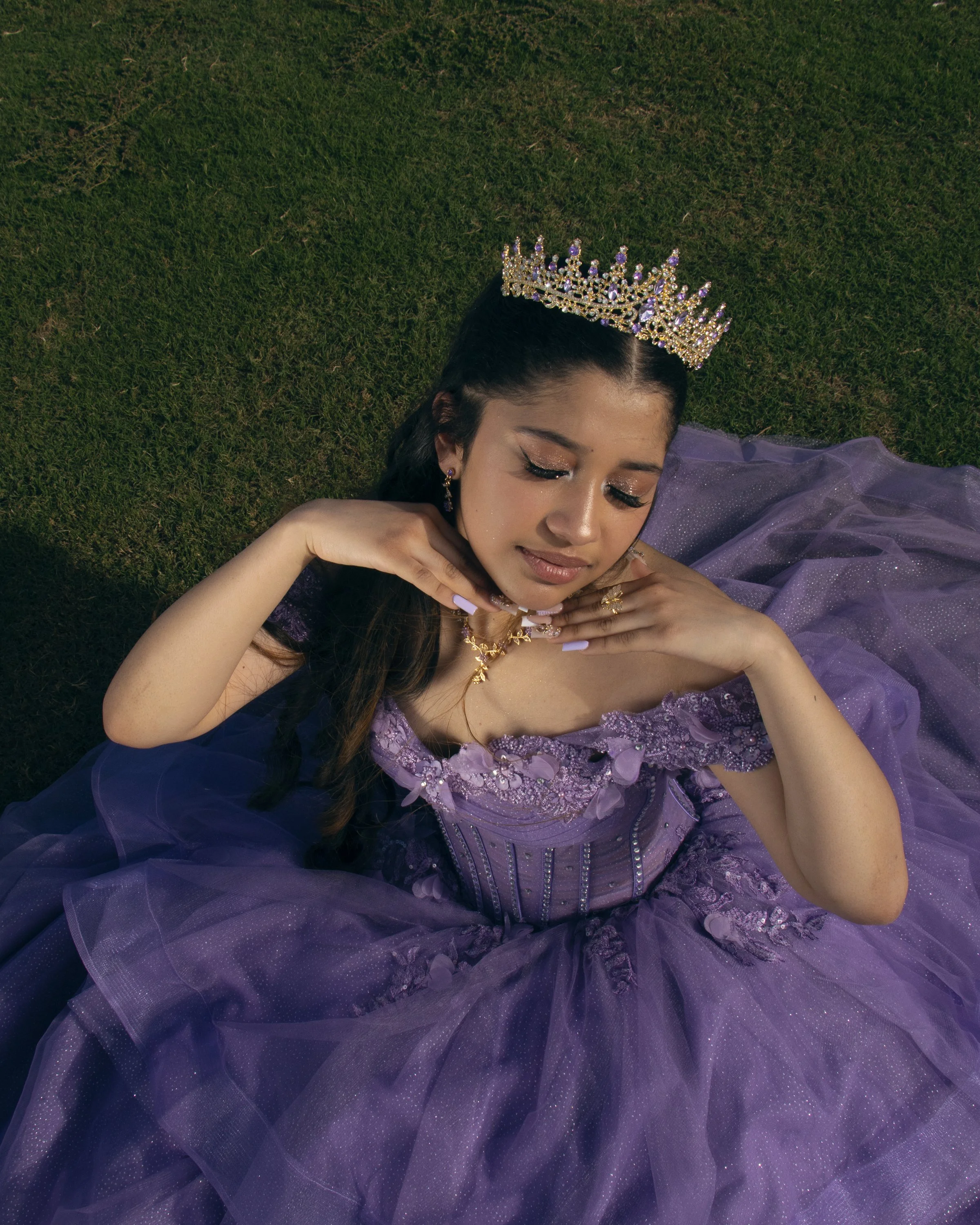 A young woman with dark hair and closed eyes lying on green grass, wearing a purple dress, golden jewelry, and a large ornate crown.
