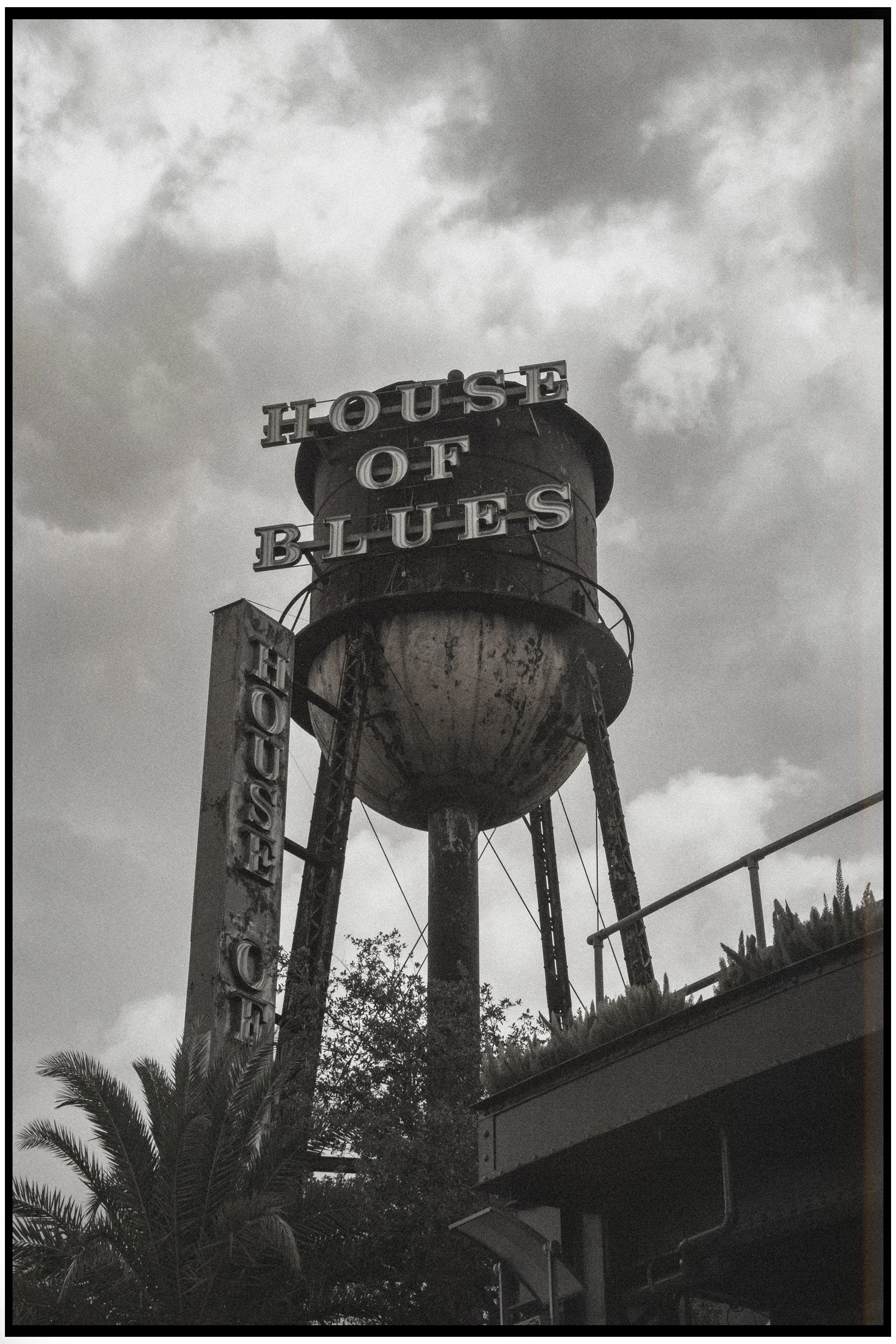 Black and white photo of an old water tower with the words 'House of Blues' on top and a vertical sign with 'Blues' on the side, trees and a building in the background, cloudy sky.