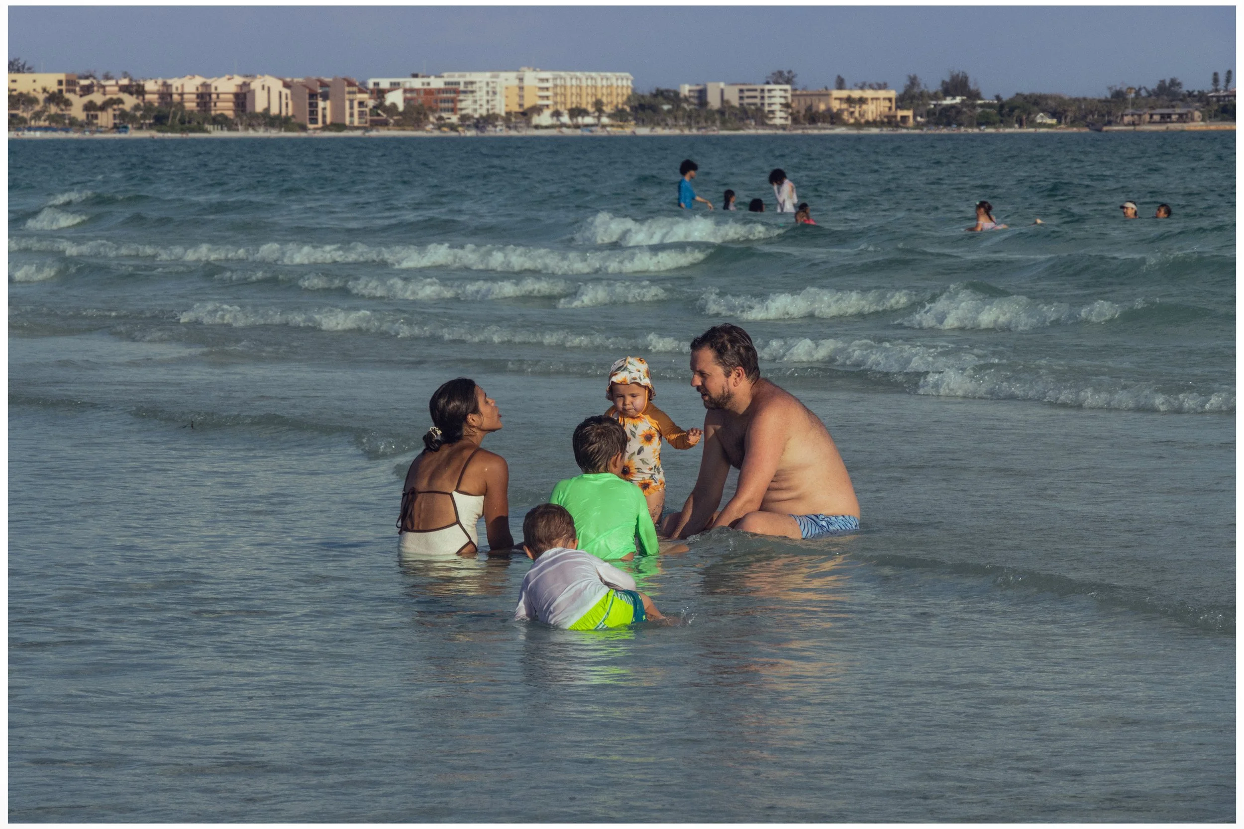 A family enjoying the beach, sitting in shallow water with children and adults, while other people swim and play further out in the ocean, and apartment buildings line the shoreline in the background.