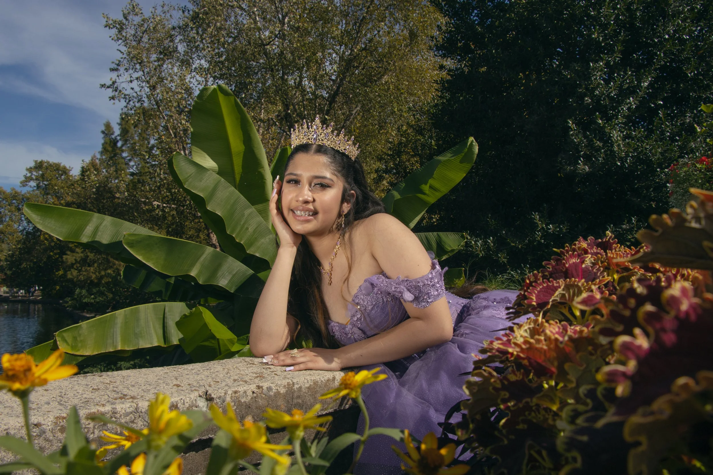 Young woman dressed as a princess with a tiara and purple gown, lying on a stone surface surrounded by colorful flowers and large green leaves, outdoors near water and trees.