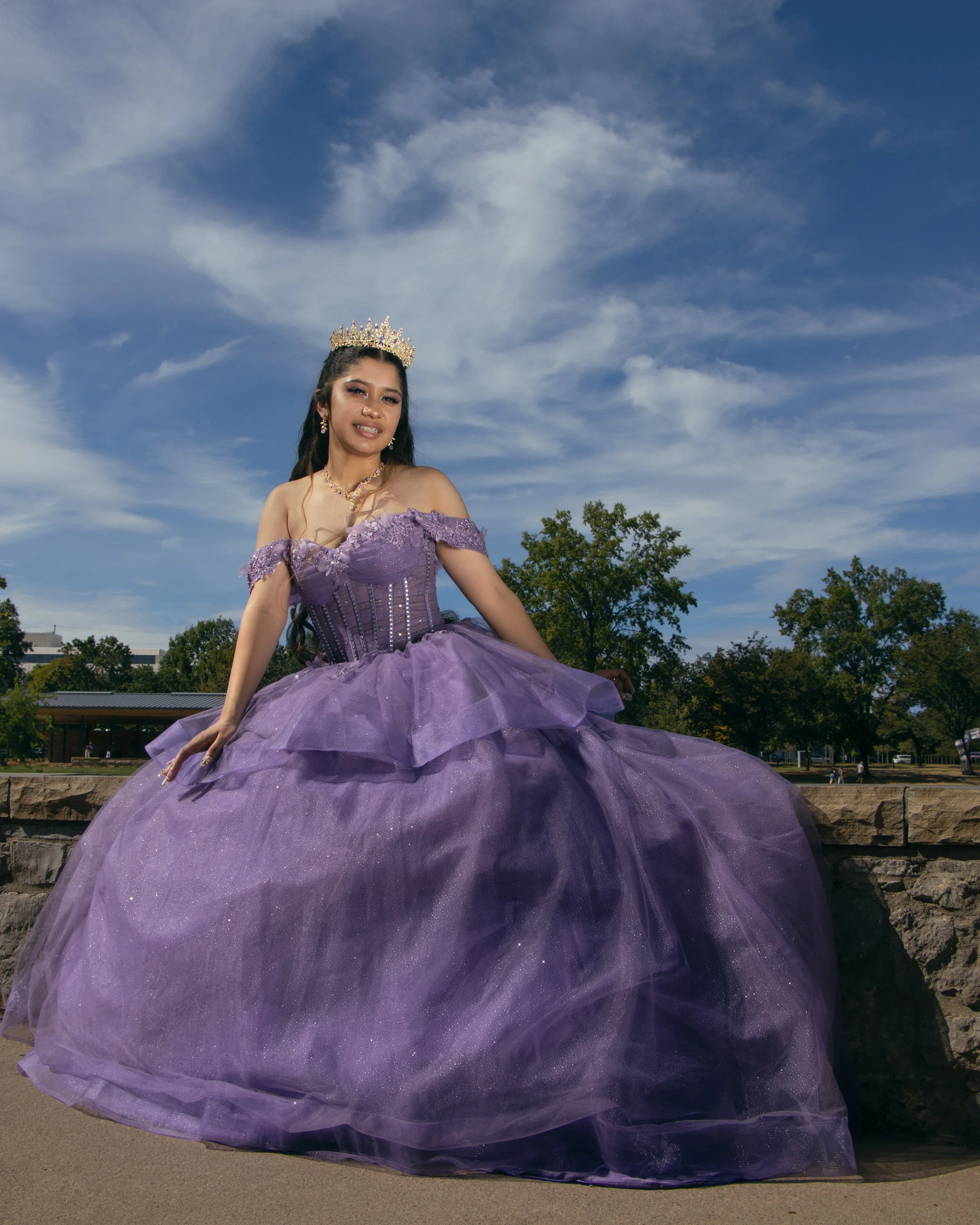 Young woman in a purple gown with off-the-shoulder lace top and a full tulle skirt, wearing a crown and jewelry, outdoors under a partly cloudy sky.