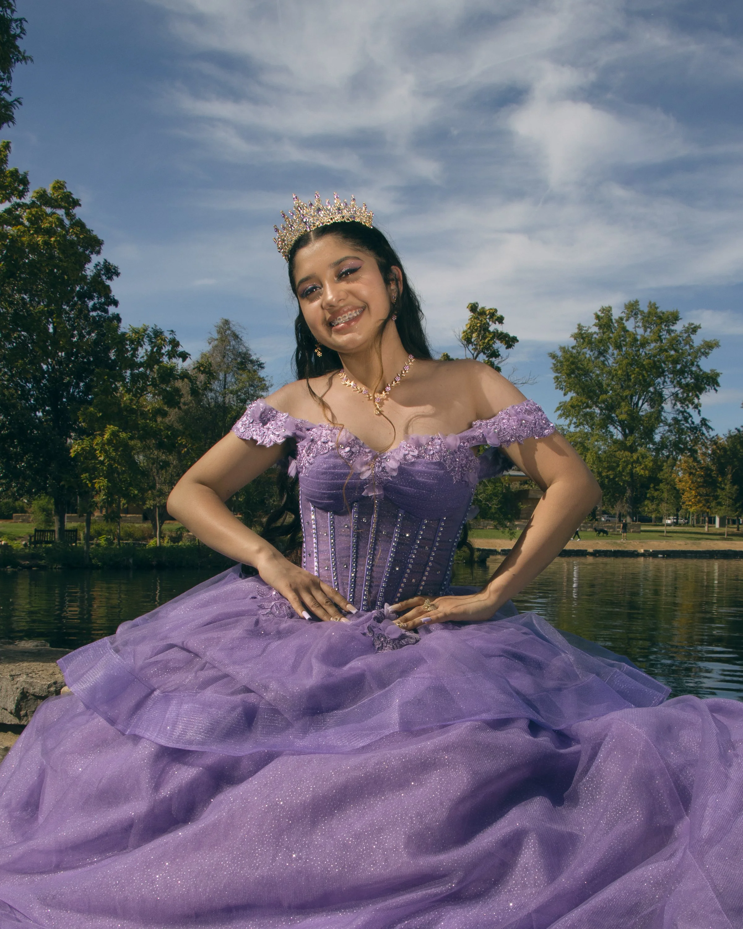 A young woman wearing a purple gown and a jeweled tiara, sitting outdoors by a body of water with trees and a park in the background.