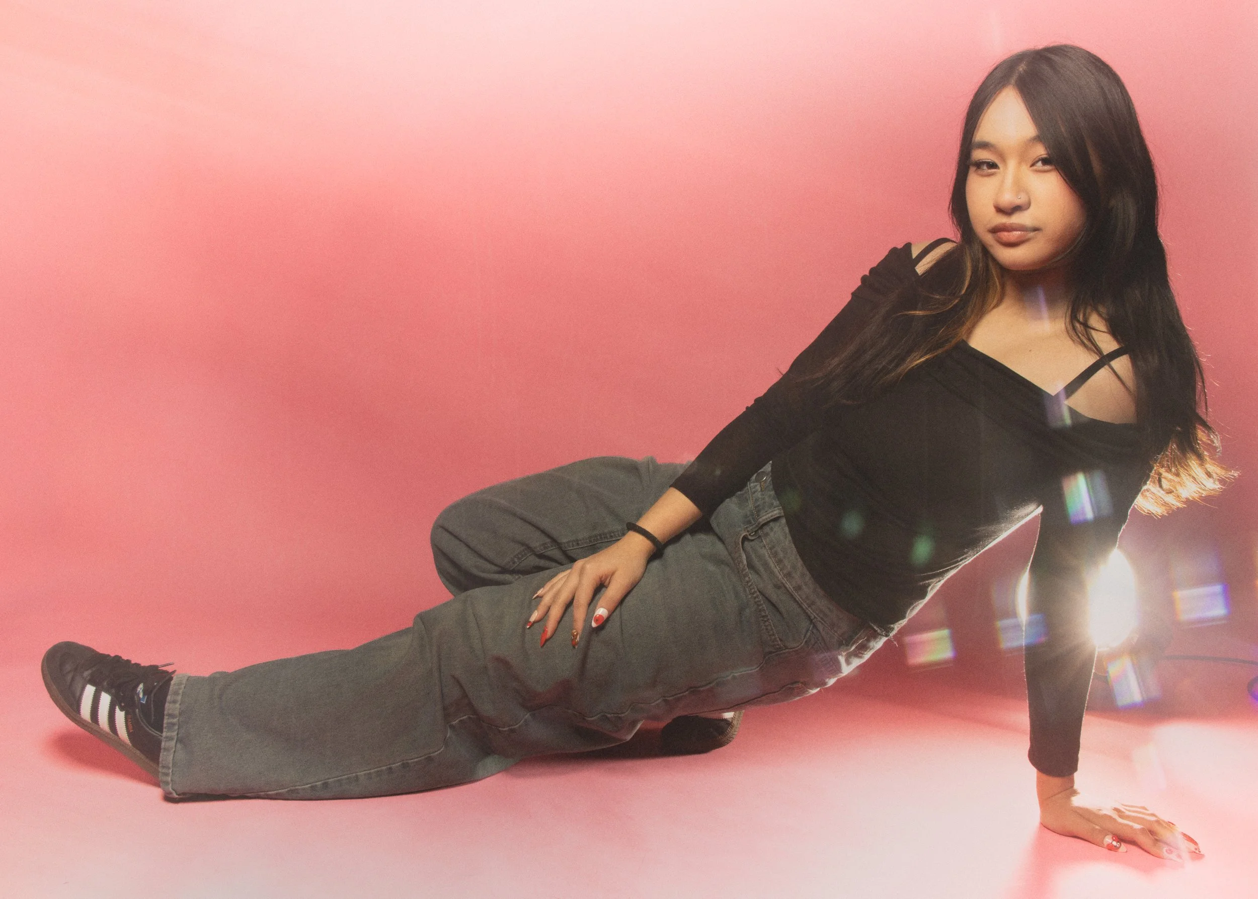 Young woman with long dark hair winking and winking, dressed in a black top and gray pants, sitting on the floor against a pink background, with lens flare effects.