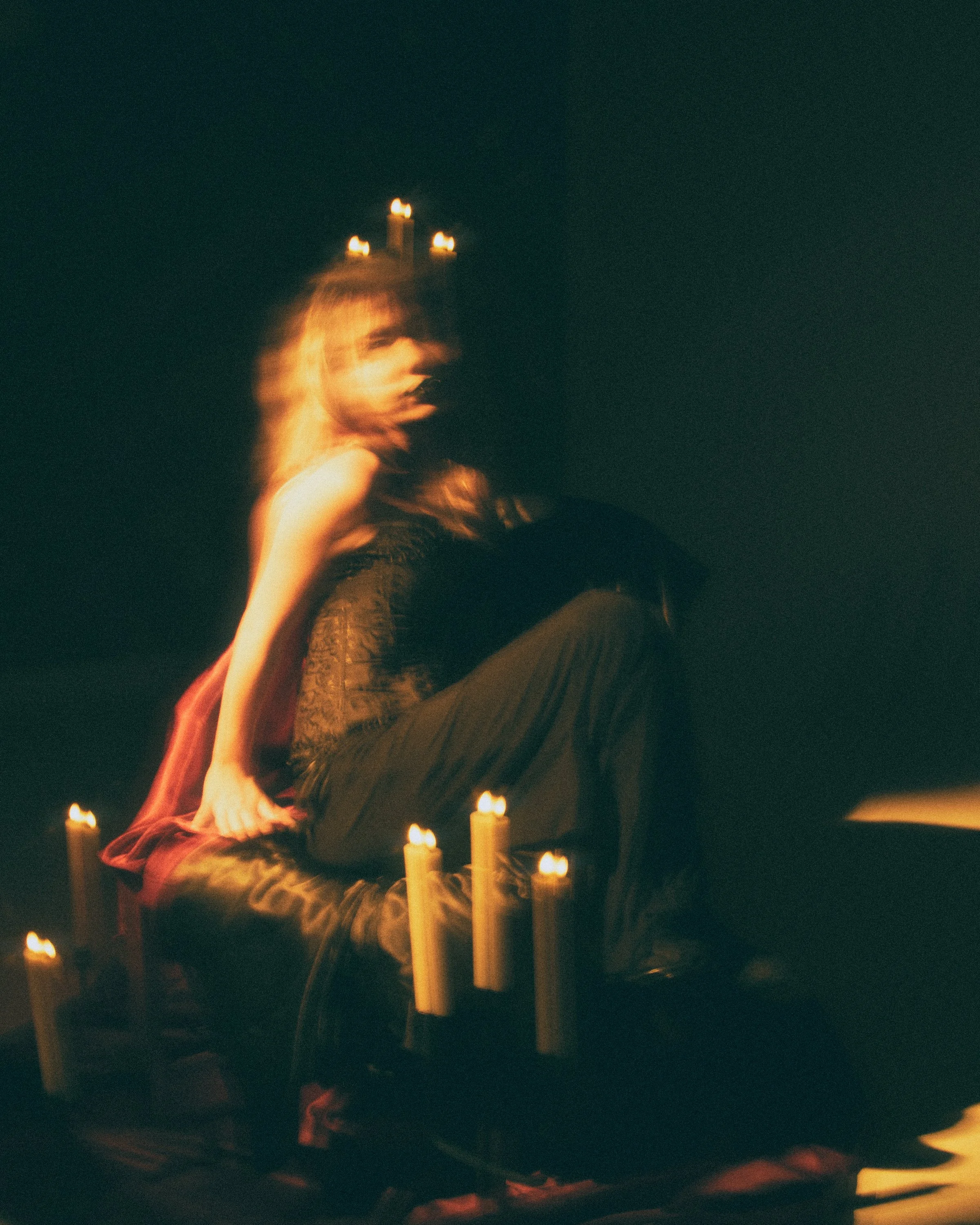 A woman with long hair and a face mask sitting on the floor surrounded by lit candles in a dark space.