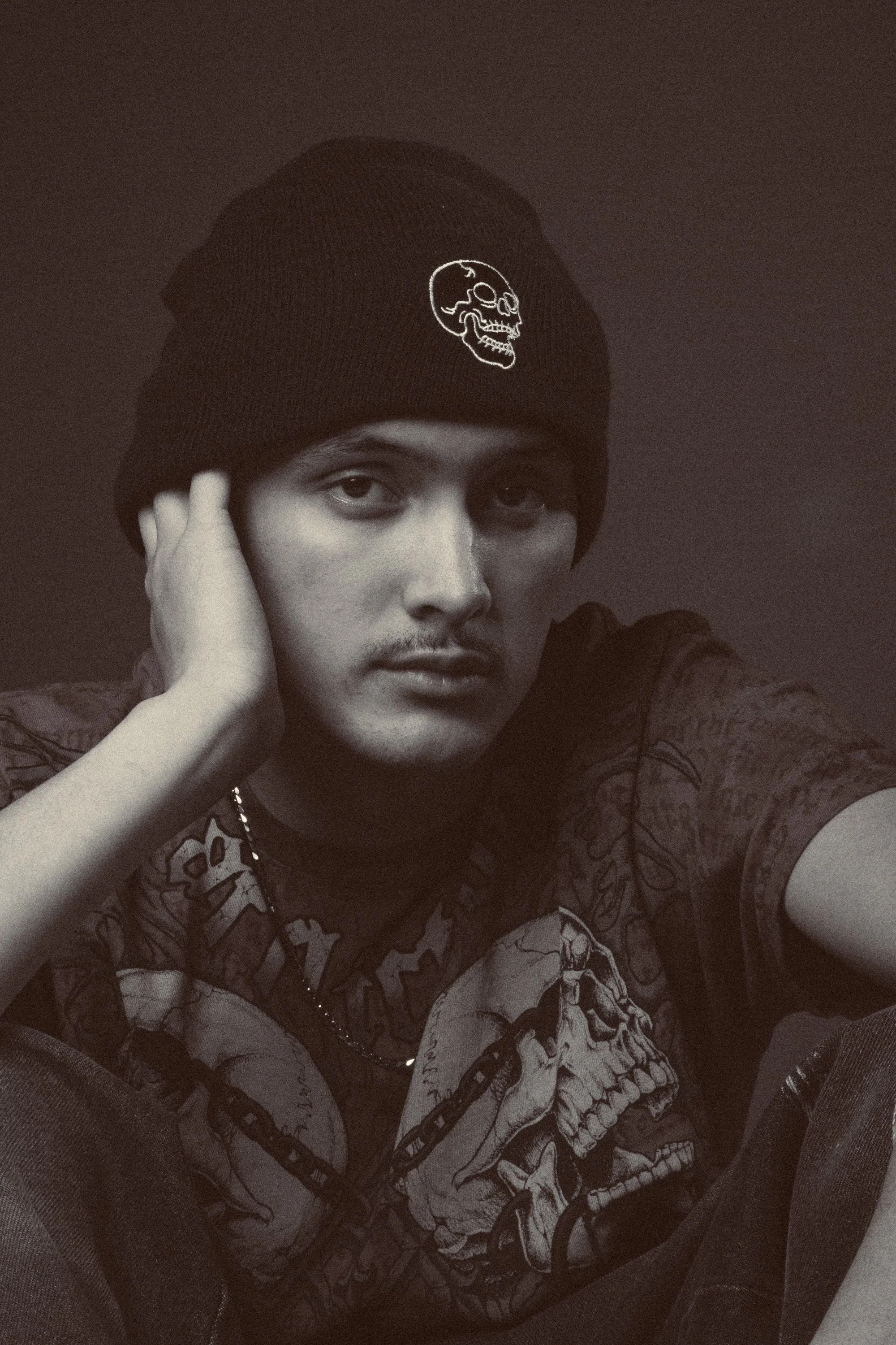 Black and white photo of a person wearing a beanie with a skull design, looking at the camera with a serious expression, resting their head on one hand, wearing a graphic t-shirt with a skull and chains design, seated in a dark room.