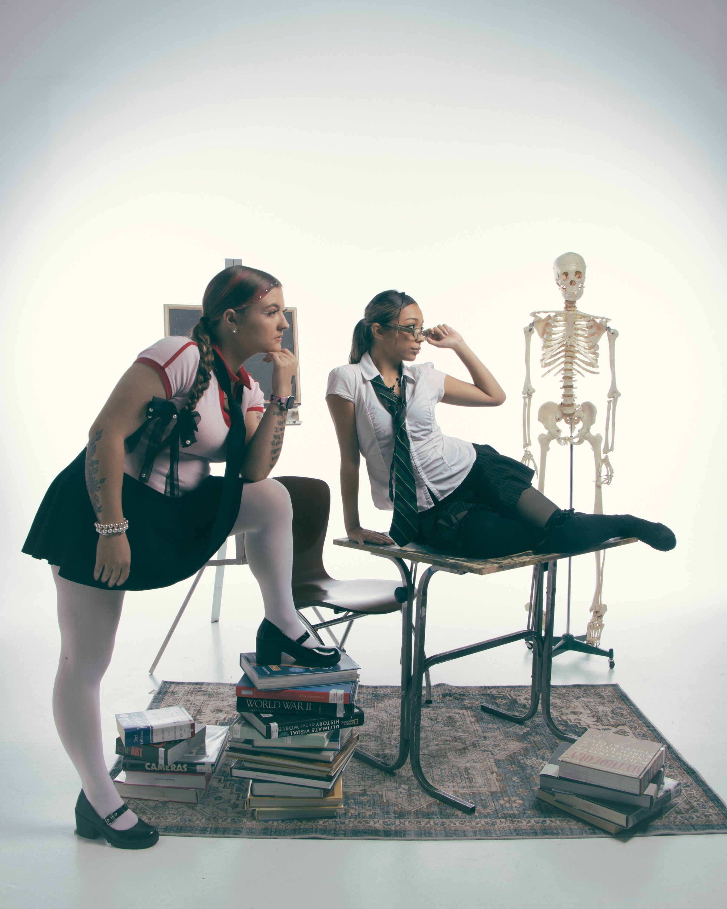 Two women in school uniforms, one with red hair and the other with dark hair, sitting on desks surrounded by stacks of books, with a human skeleton model nearby, in a room with a white background.