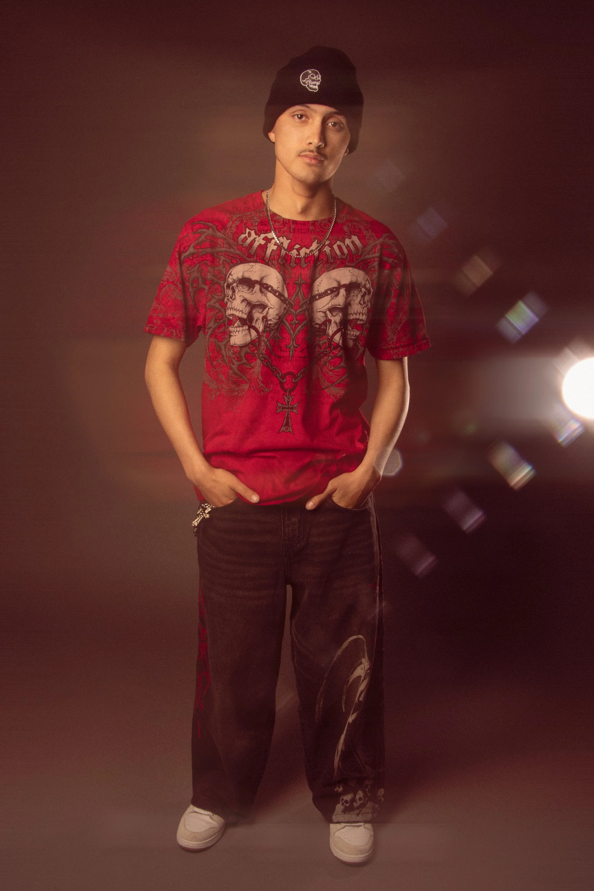 Young man standing against a dark background, wearing a black beanie with a skull logo, a red graphic T-shirt with skull and chain designs, black pants with red stripe details and a skull design, white sneakers, and a silver chain necklace.