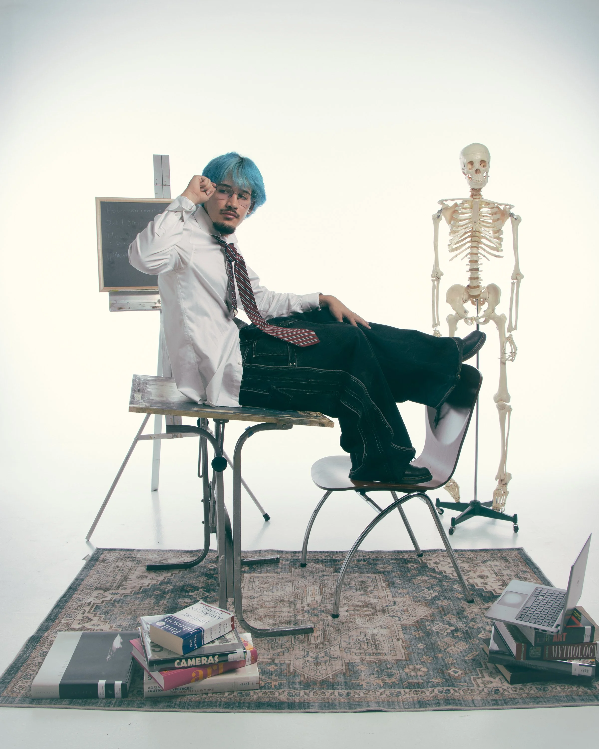 A person with blue hair and glasses sitting on a chair with his legs extended on a table, near a skeleton model and a blackboard, with books and a box labeled "Cameras" and "Mythology" on the rug and table in a bright, minimalistic room.