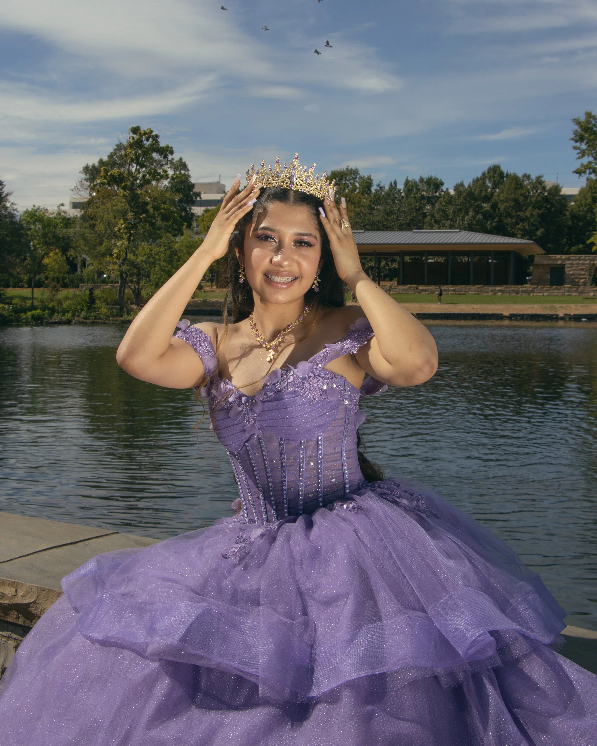 A young woman in a purple formal gown with lace details, wearing a tiara, earrings, necklace, and ring, poses outdoors near a body of water with trees and a building in the background, and a few airplanes flying overhead.