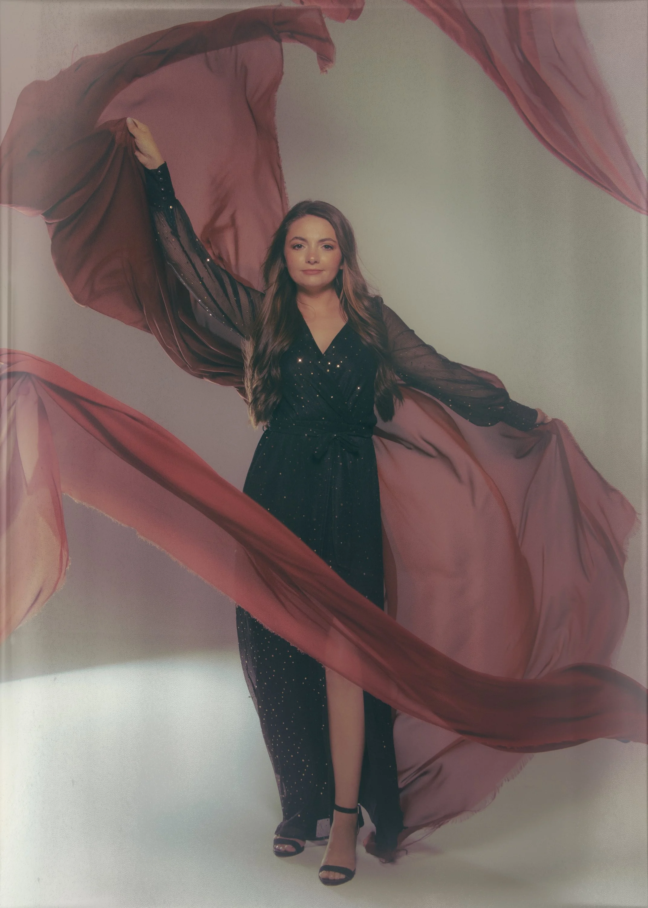 A woman with long brown hair in a black dress with sparkles, black ankle-strap high heels, and black socks, stands with one arm raised, holding a flowing, sheer pink fabric that billows around her against a plain background.