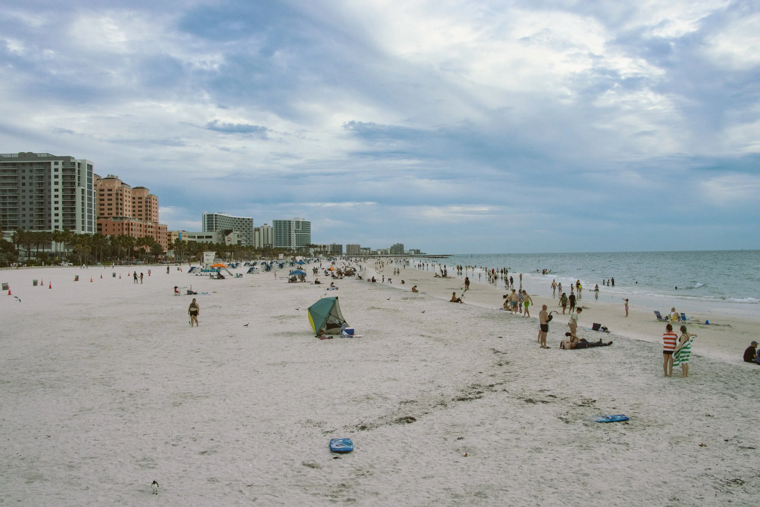 A busy beach scene with many people relaxing and swimming. Tall buildings and hotels line the background along the shoreline, under a partly cloudy sky.