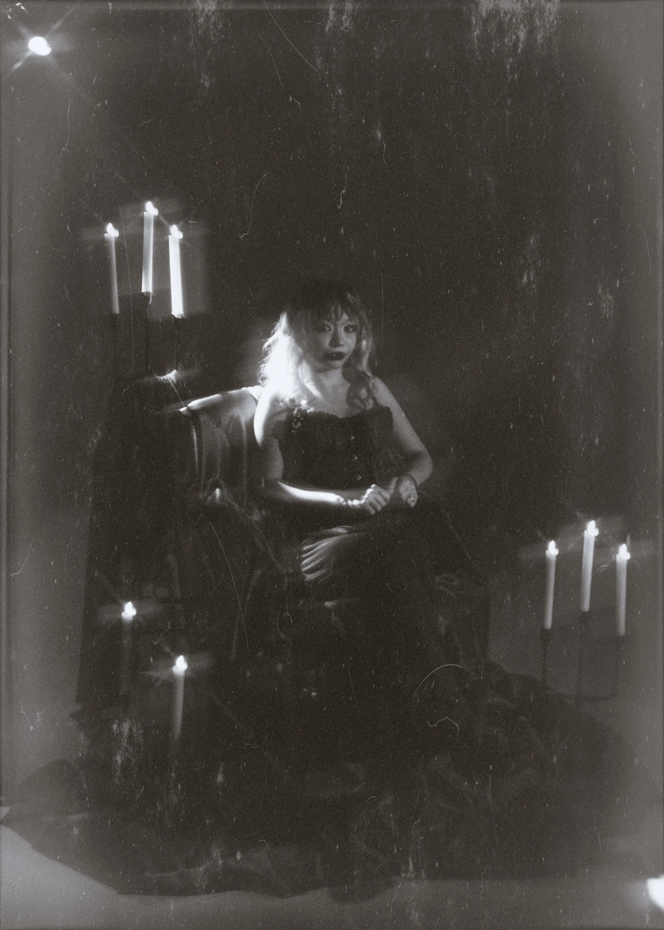 Black and white photo of a woman with wavy hair, Sitting on a dark chair, with candles around her, in a dimly lit room with a dark background.
