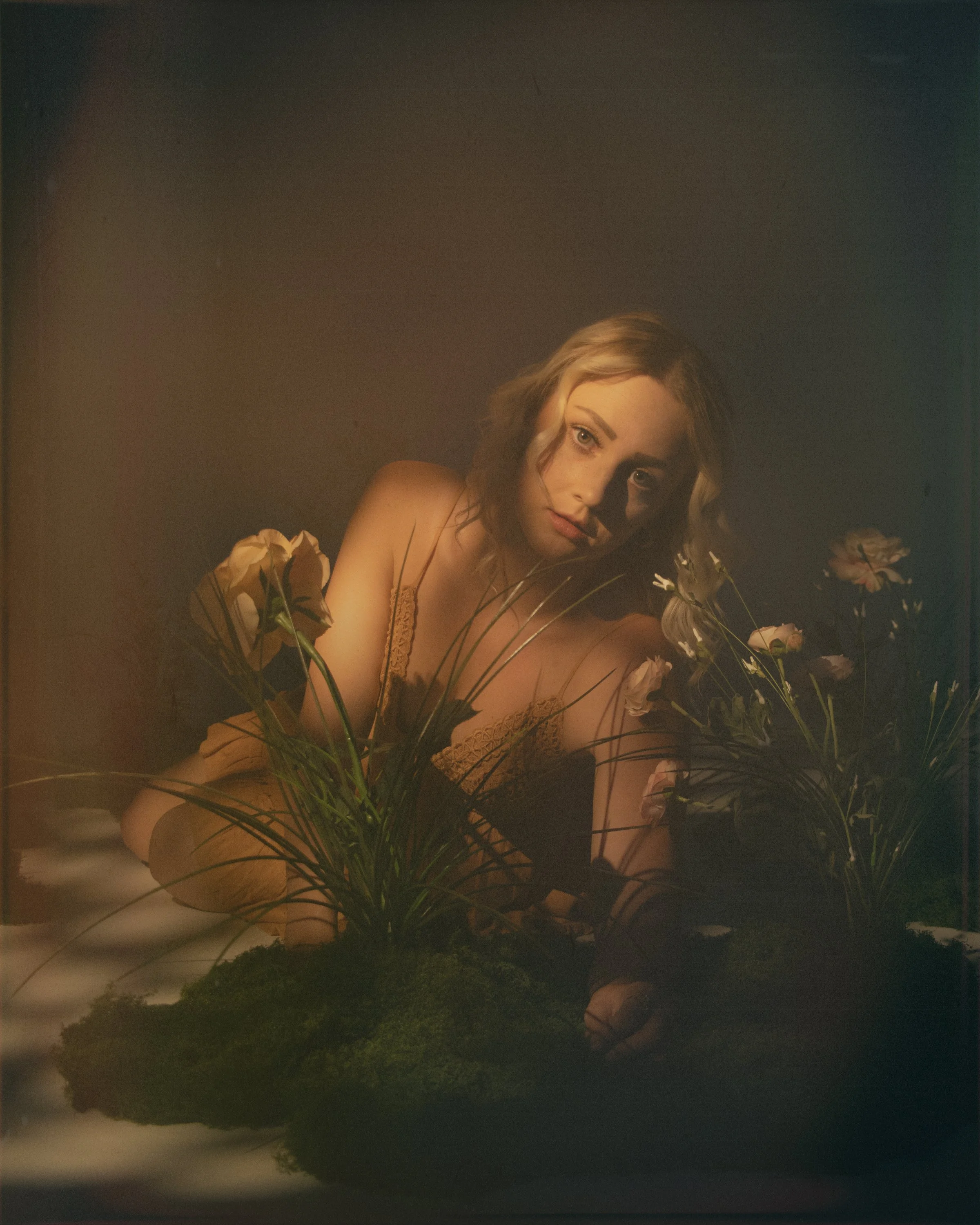 A young woman with long blonde wavy hair, wearing a beige lace dress, is sitting behind a cluster of pink and white flowers, with a dark background and soft warm lighting highlighting her face.