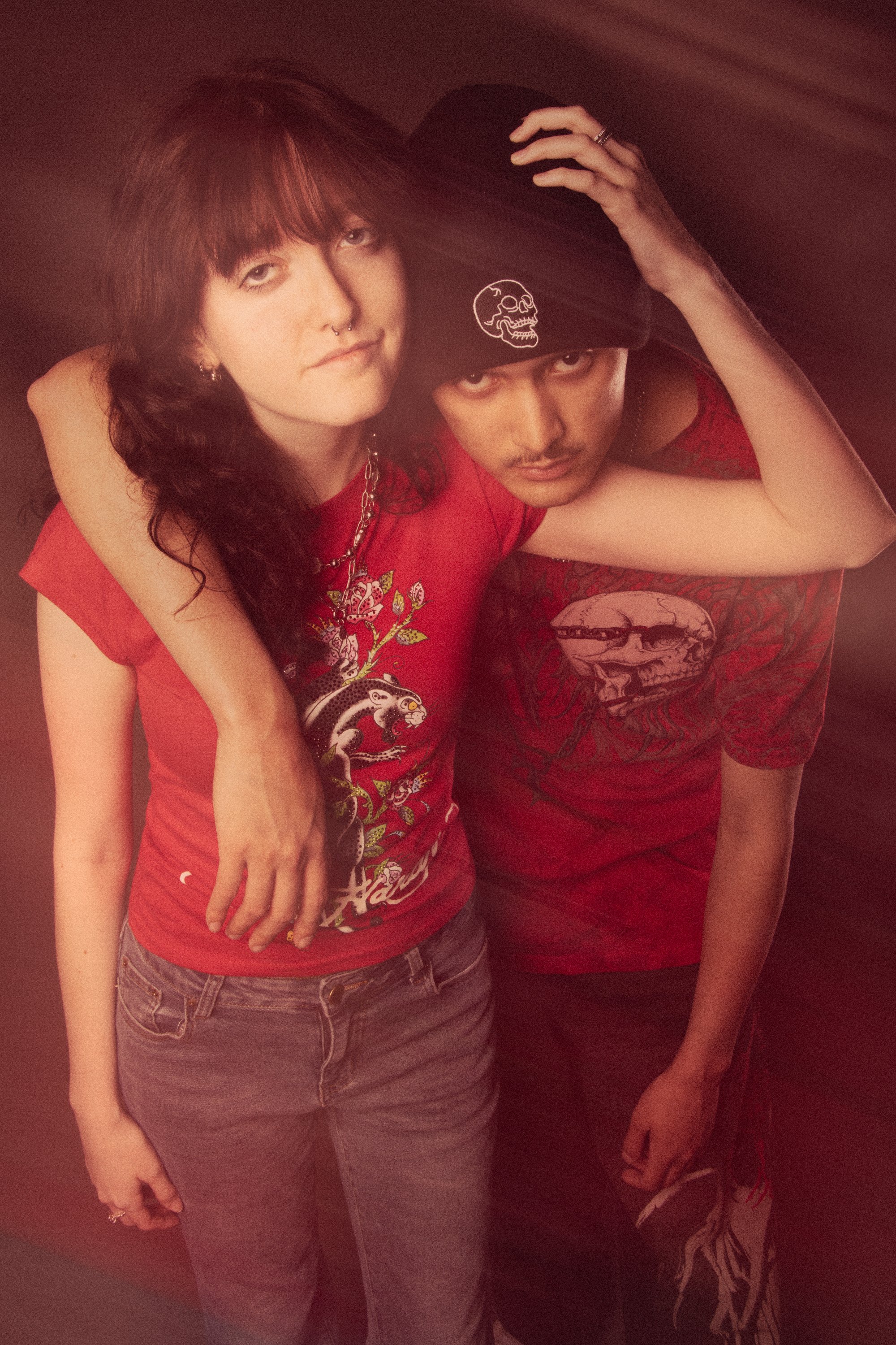 A young woman with red hair and a septum piercing wearing a red graphic t-shirt and gray jeans, standing next to a person with a darker complexion wearing a black beanie with a skull logo, a red graphic shirt, and patterned pants. They are posed clos