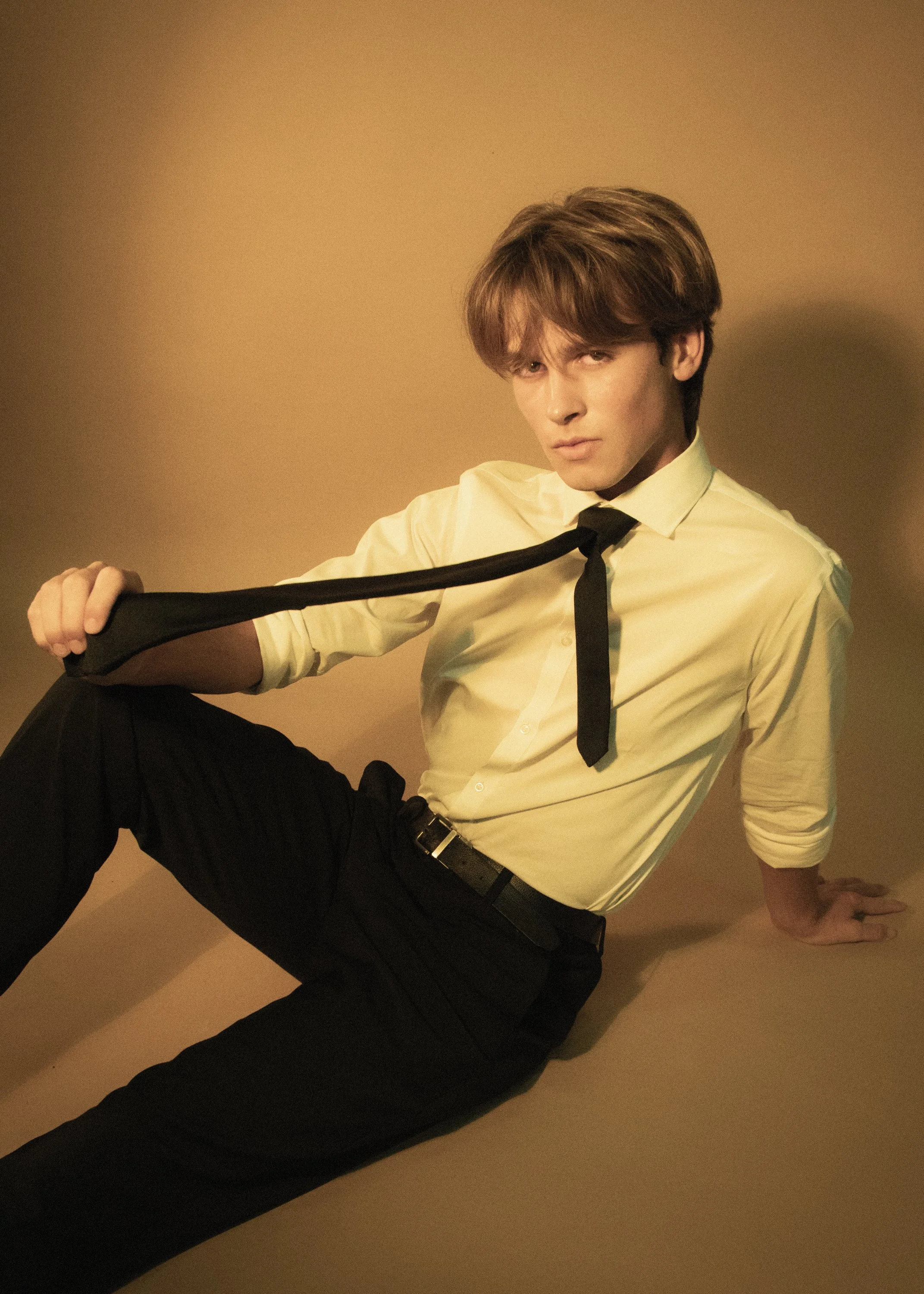A young man with brown hair wearing a light yellow dress shirt, black tie, and black pants, sitting on the floor against a plain background, looking at the camera with a serious expression.