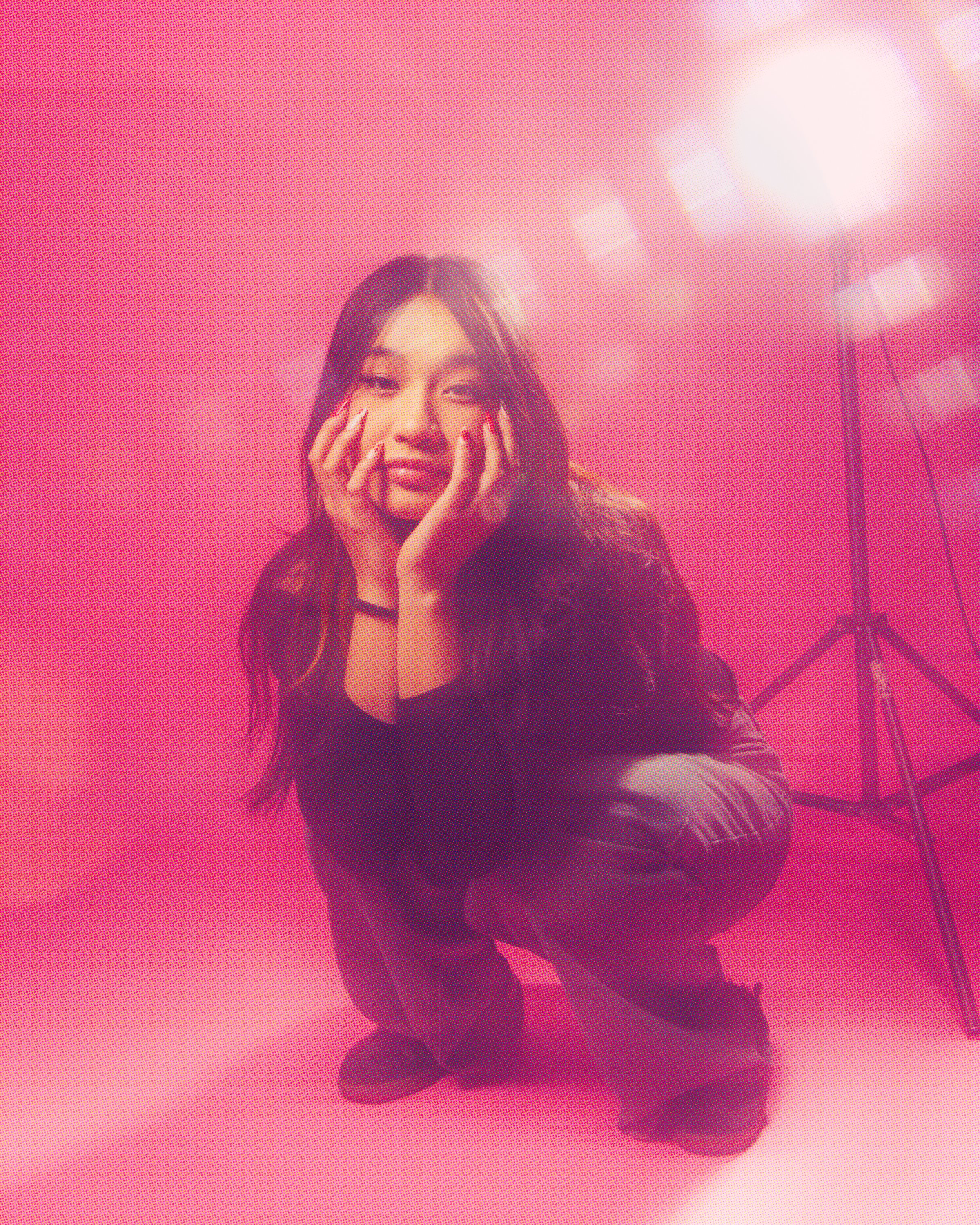 A woman posing in front of a bright pink background, with her hands on her face and a slight smile, wearing a black outfit.