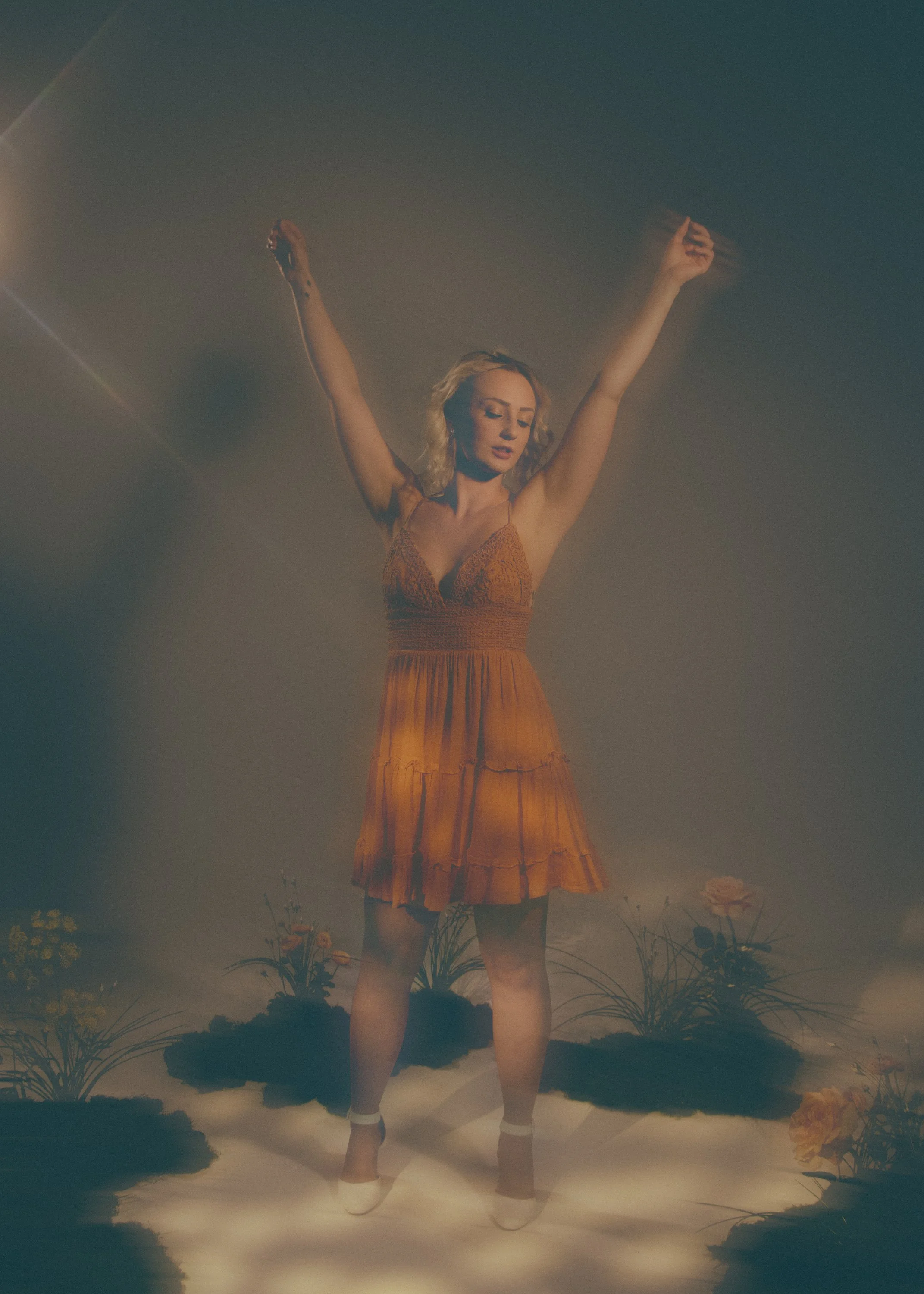 A woman in an orange dress with her arms raised stands amidst flowers and greenery, with a dreamy misty atmosphere and clouds or fog at her feet.