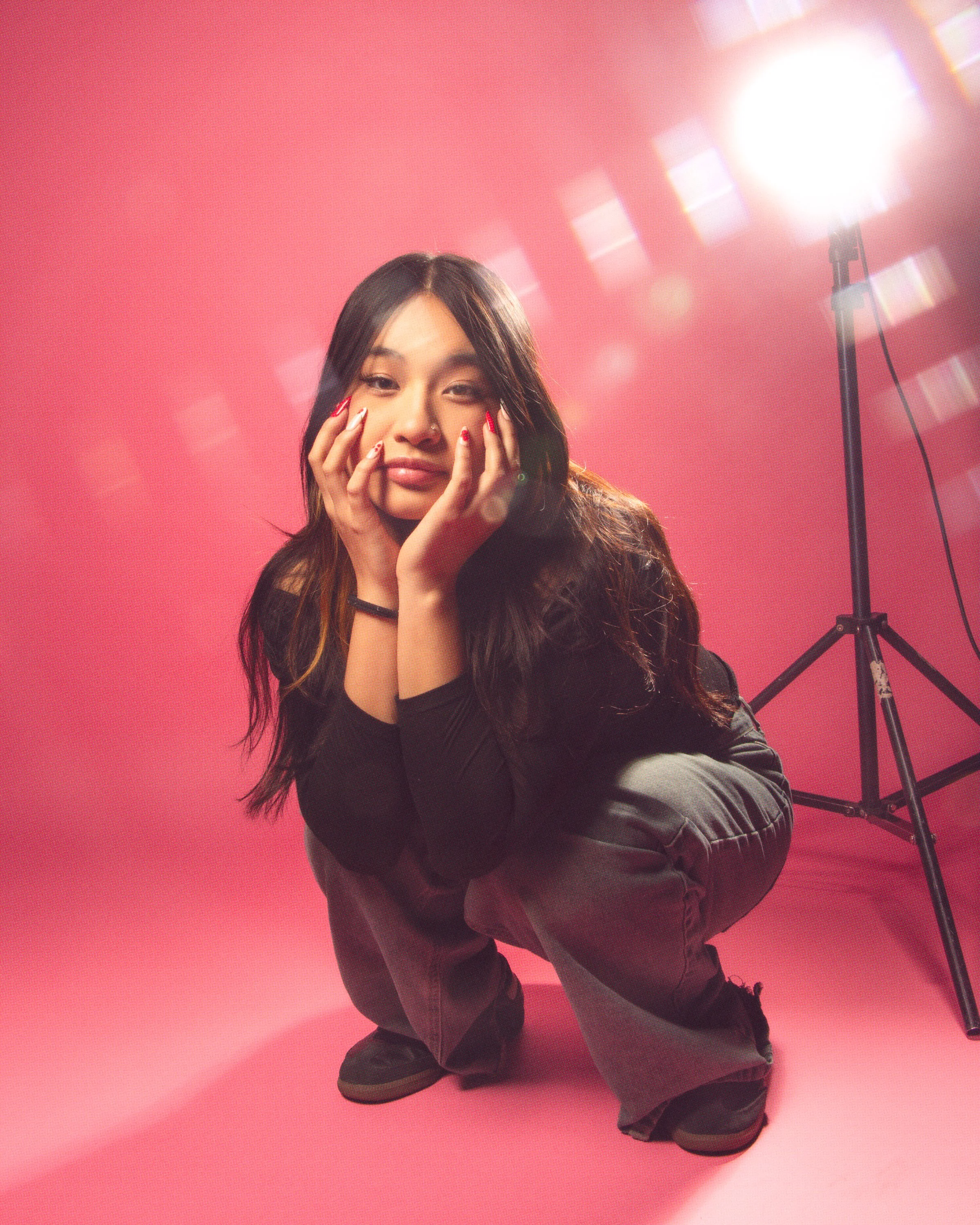 A woman with black hair and red nail polish crouching on a pink floor, with bright studio lighting behind her.