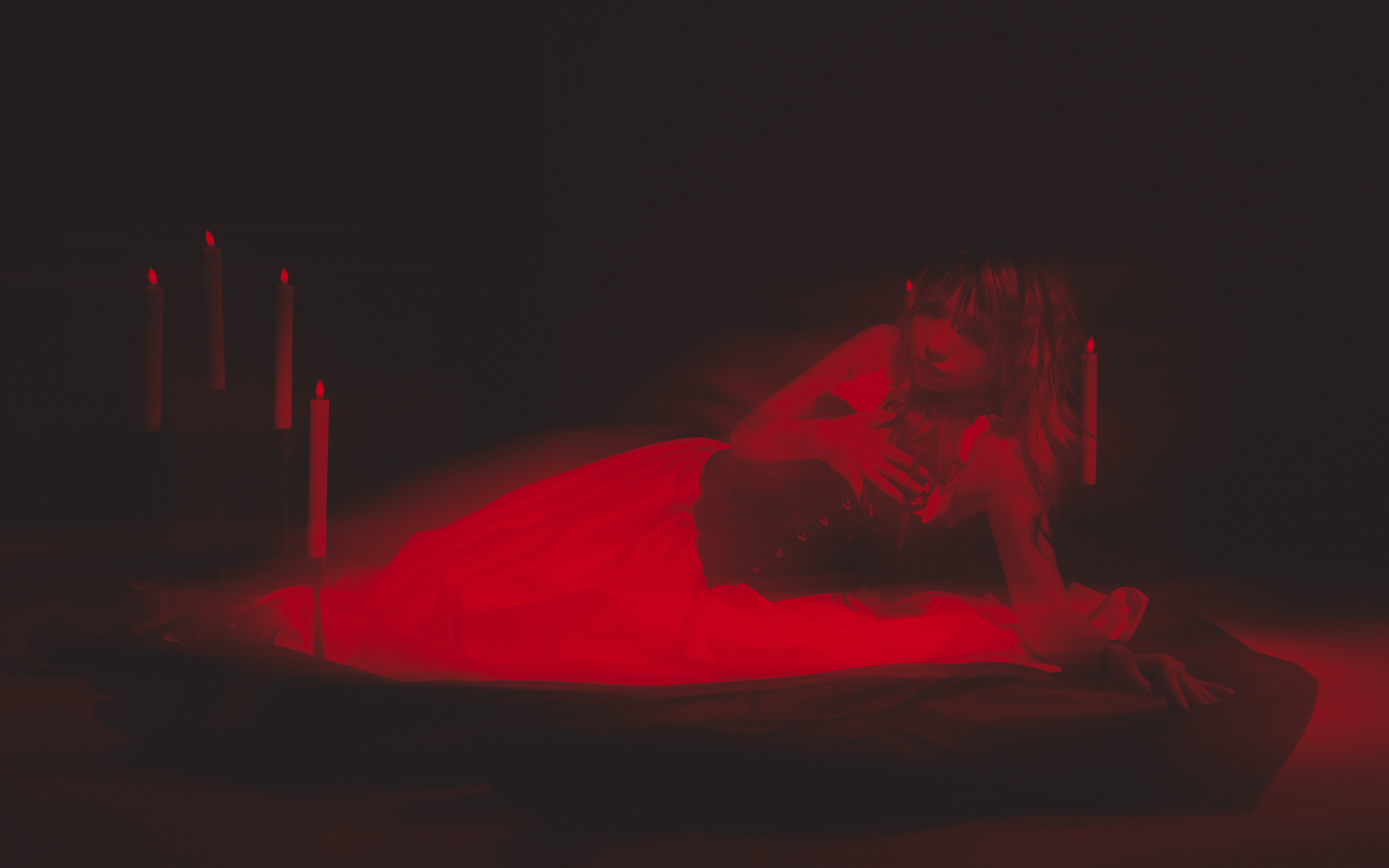 A woman with long wavy hair dressed in a red gown, lying on her side on the floor in a dark room with red lighting, surrounded by tall red candles.