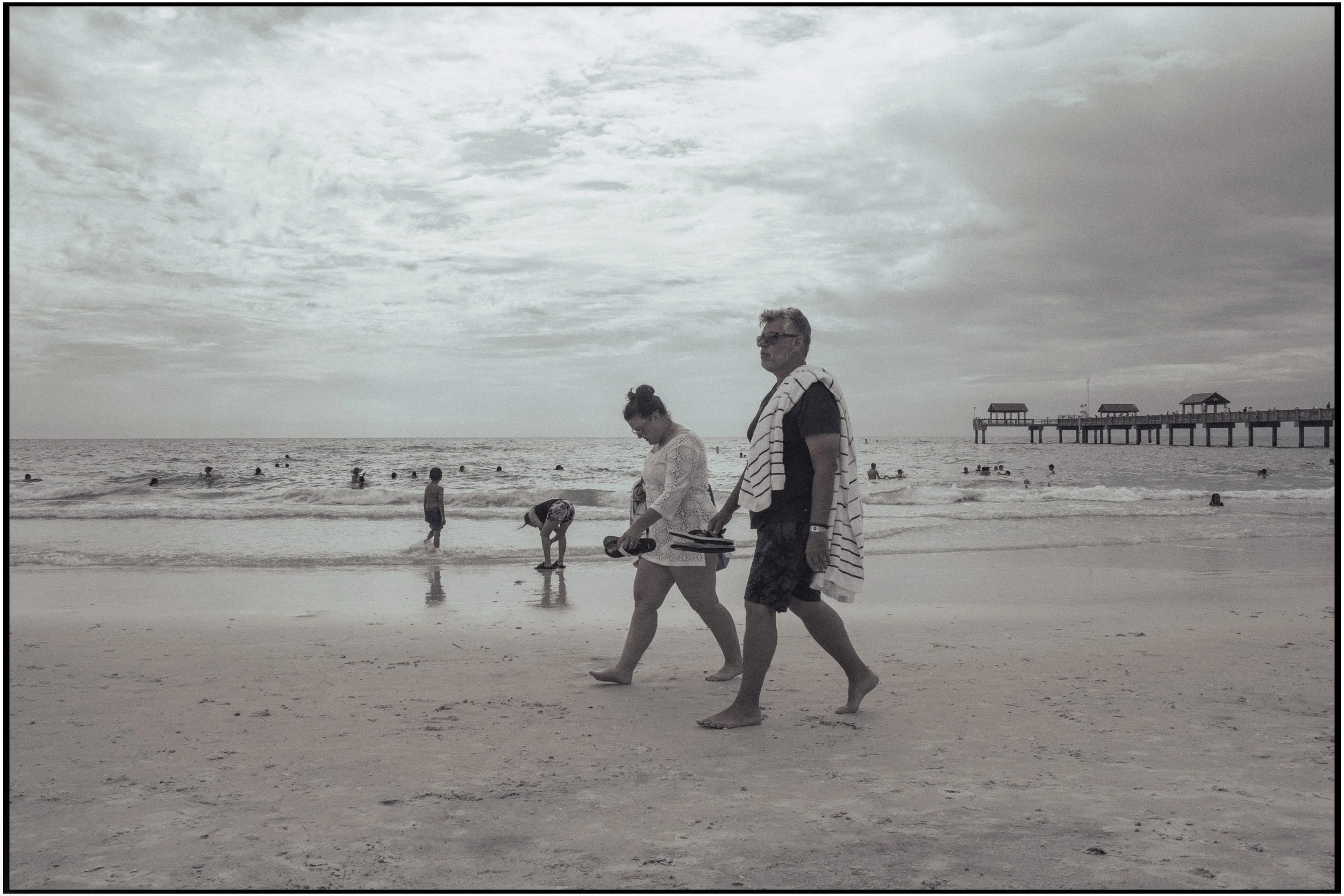 People walking along a beach with many people swimming in the ocean. There is a pier with small pavilions extending into the water in the background. The sky is cloudy.