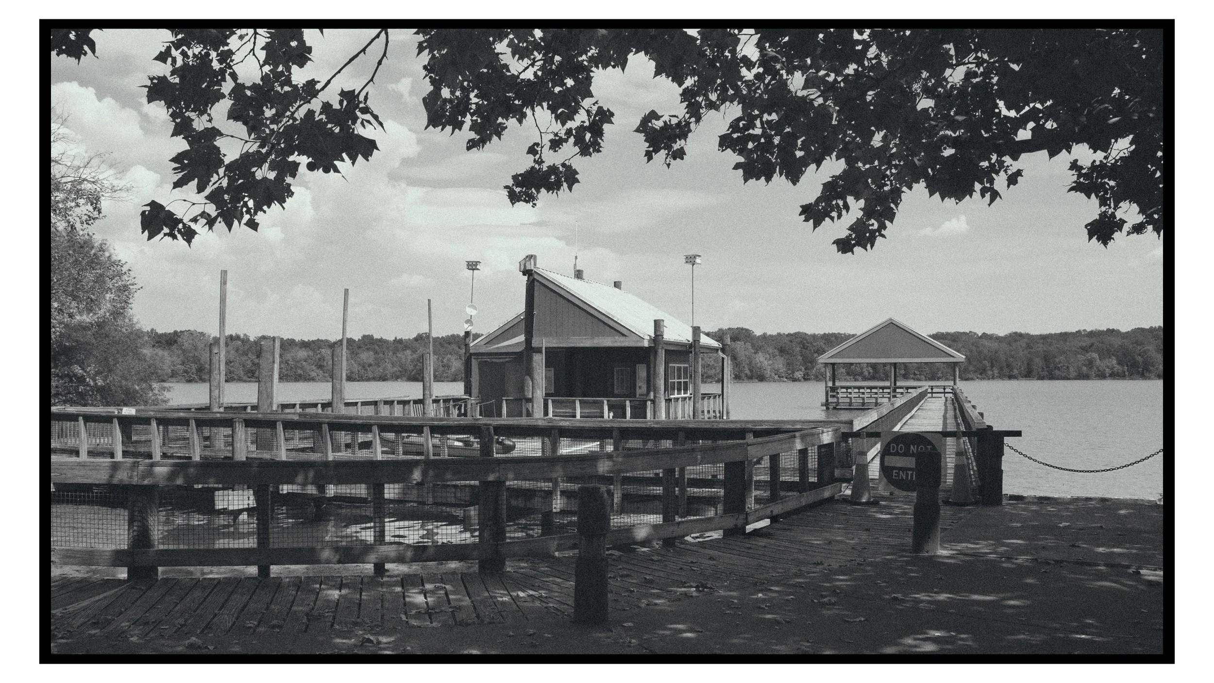 A black and white photo of a lakeside dock with a small building and a covered pier, surrounded by trees and water, and a cloudy sky.