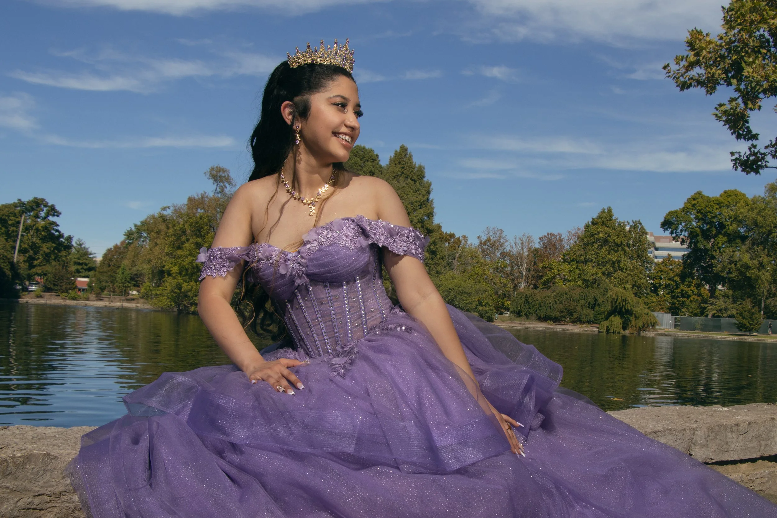 A young woman dressed in a purple ball gown with off-the-shoulder lace details and jewelry, sitting outdoors by a body of water with trees and a blue sky in the background.