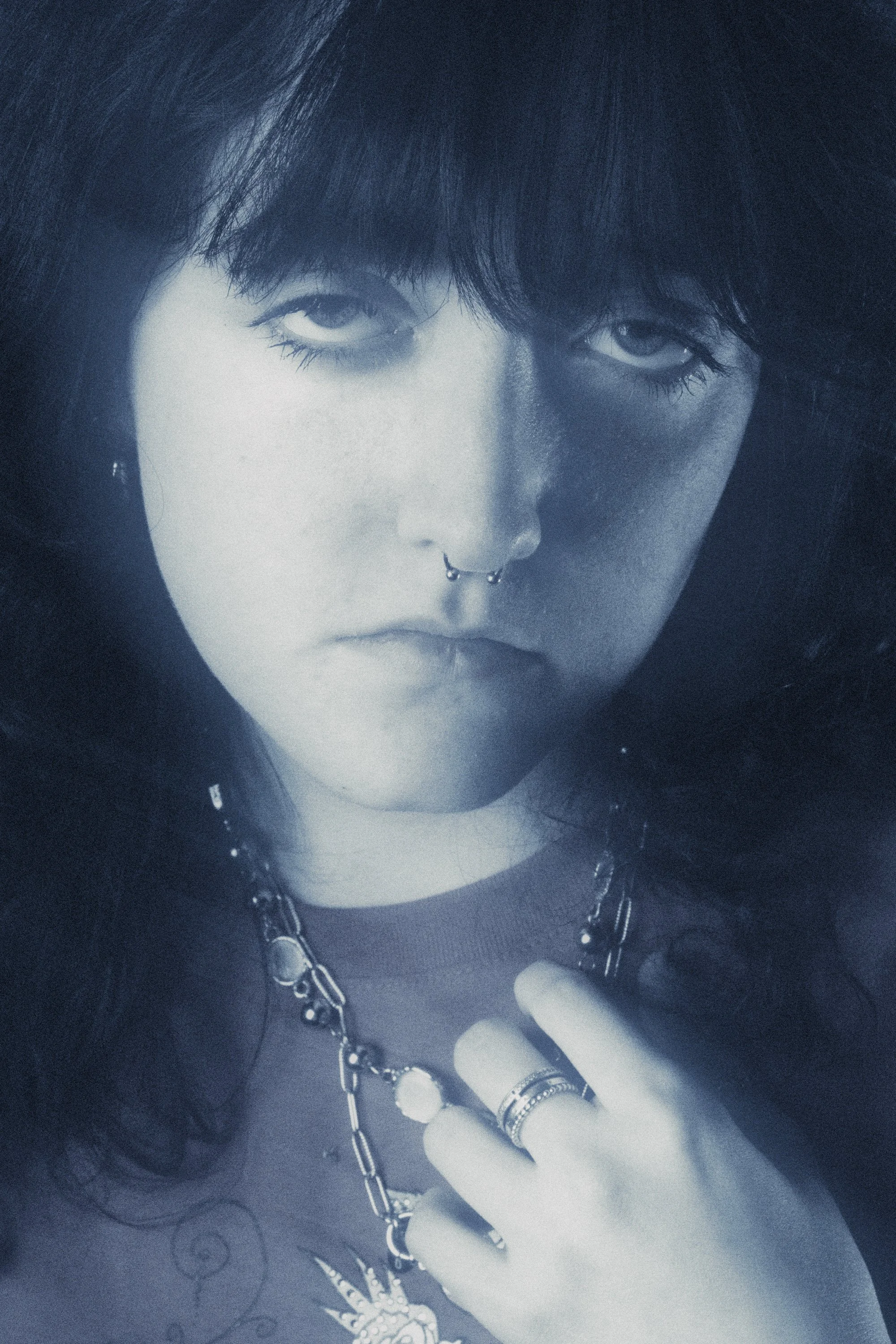 A black and white close-up portrait of a woman with dark hair and bangs, wearing layered necklaces, rings, and a septum piercing, with a serious expression and hand near her face.