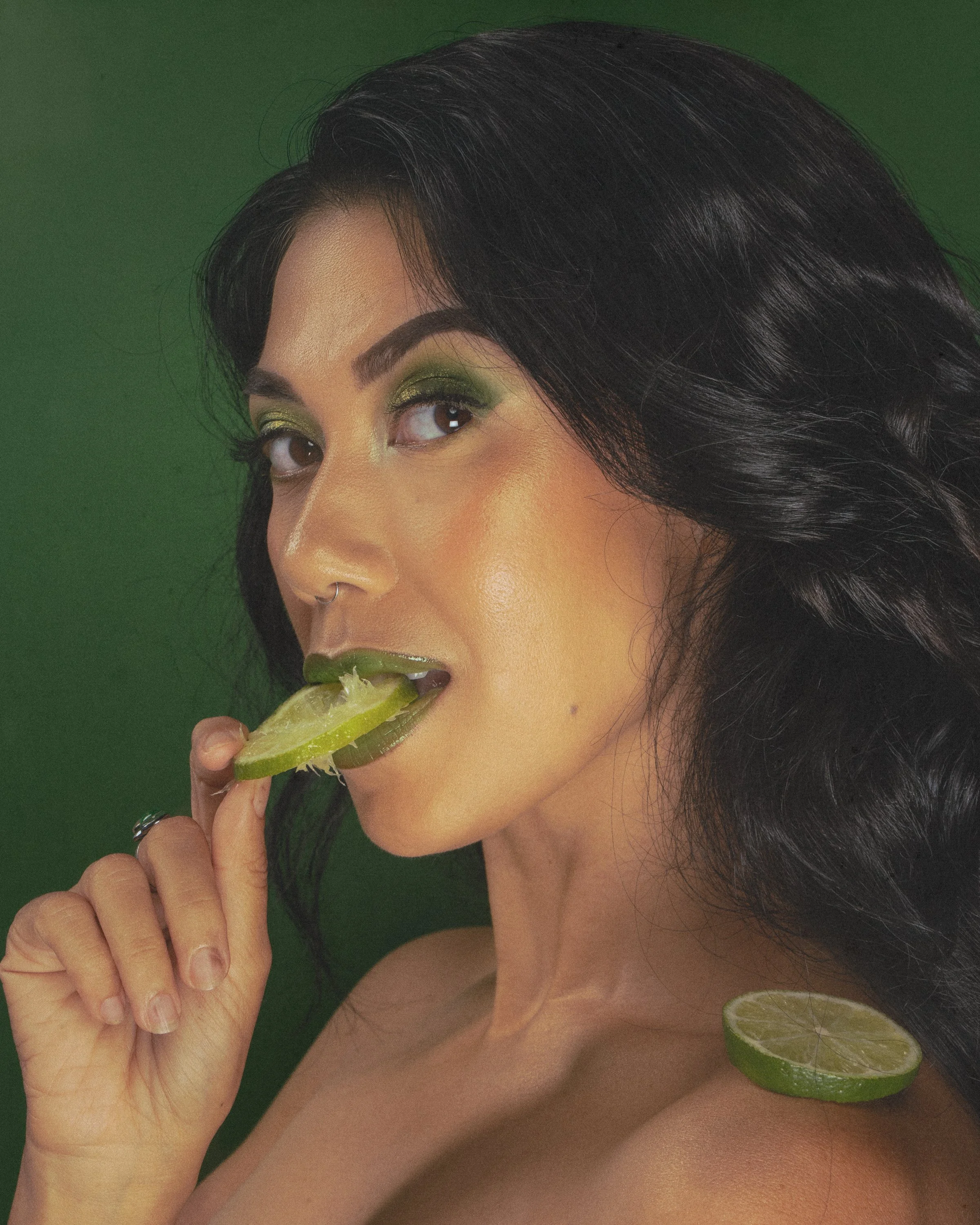 Close-up of a woman with long black hair, green eye makeup, and a nose ring, holding a slice of lime near her mouth, with lime slices on her shoulder and chest against a green background.