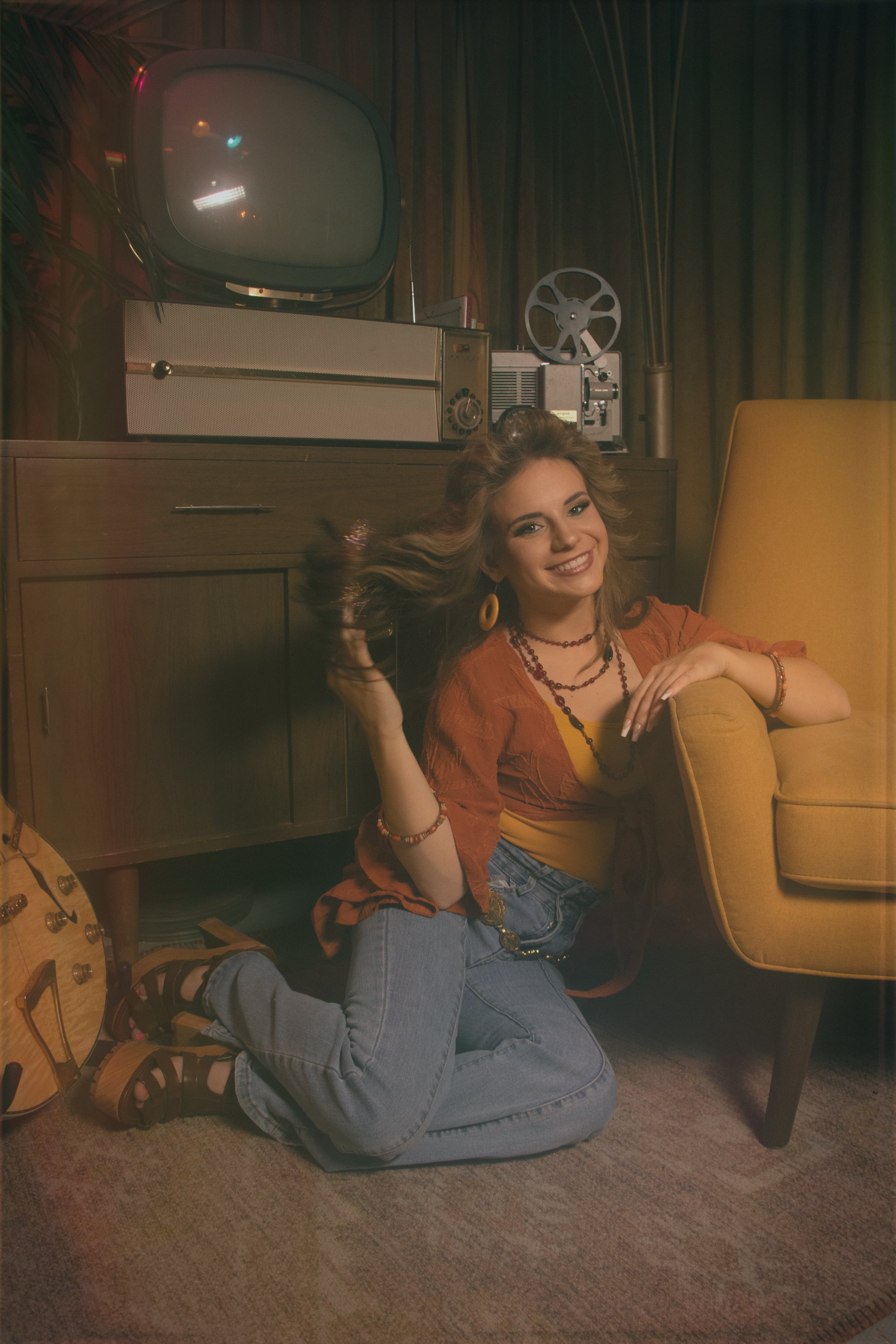 A smiling woman with blonde hair sitting on her knees on a carpeted floor, leaning against a yellow armchair. She is wearing a yellow top, a red open shirt, blue jeans, and black sandals. Behind her, there is a wooden cabinet with an old-fashioned te