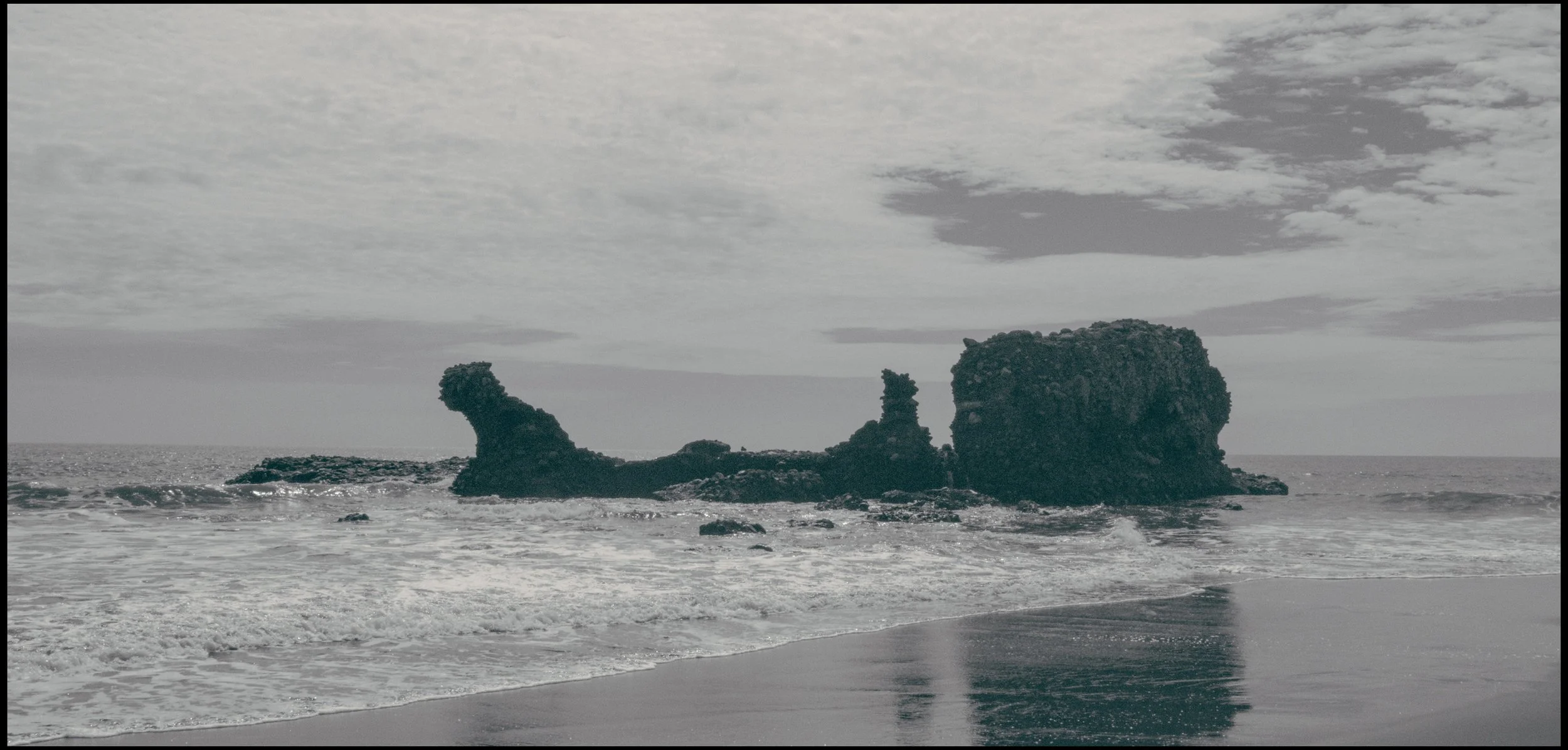 Black-and-white photograph of ocean waves crashing on a sandy beach with three large rocky formations emerging from the water.
