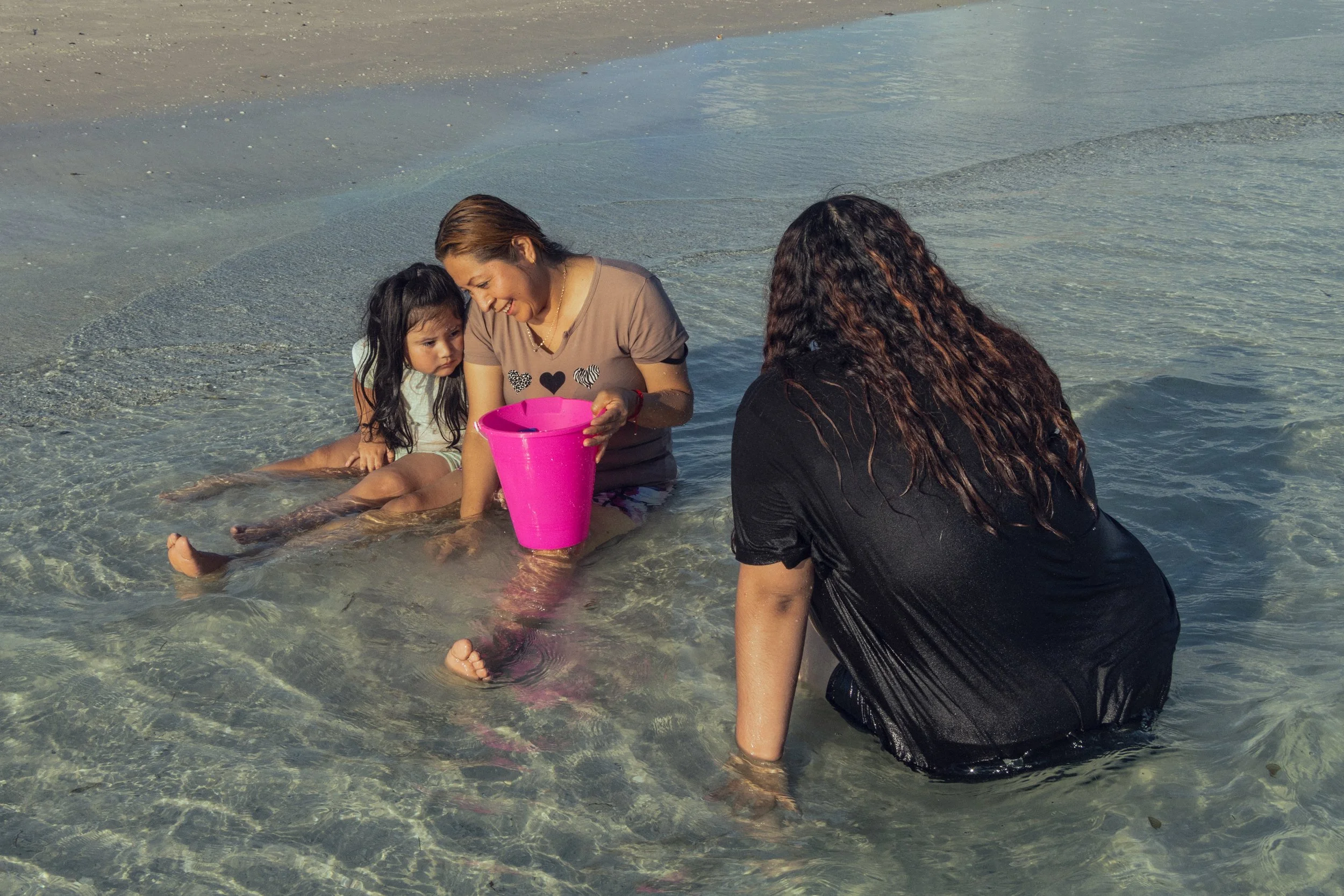Three women and a girl sitting in shallow water at the beach, with one woman holding a pink bucket.