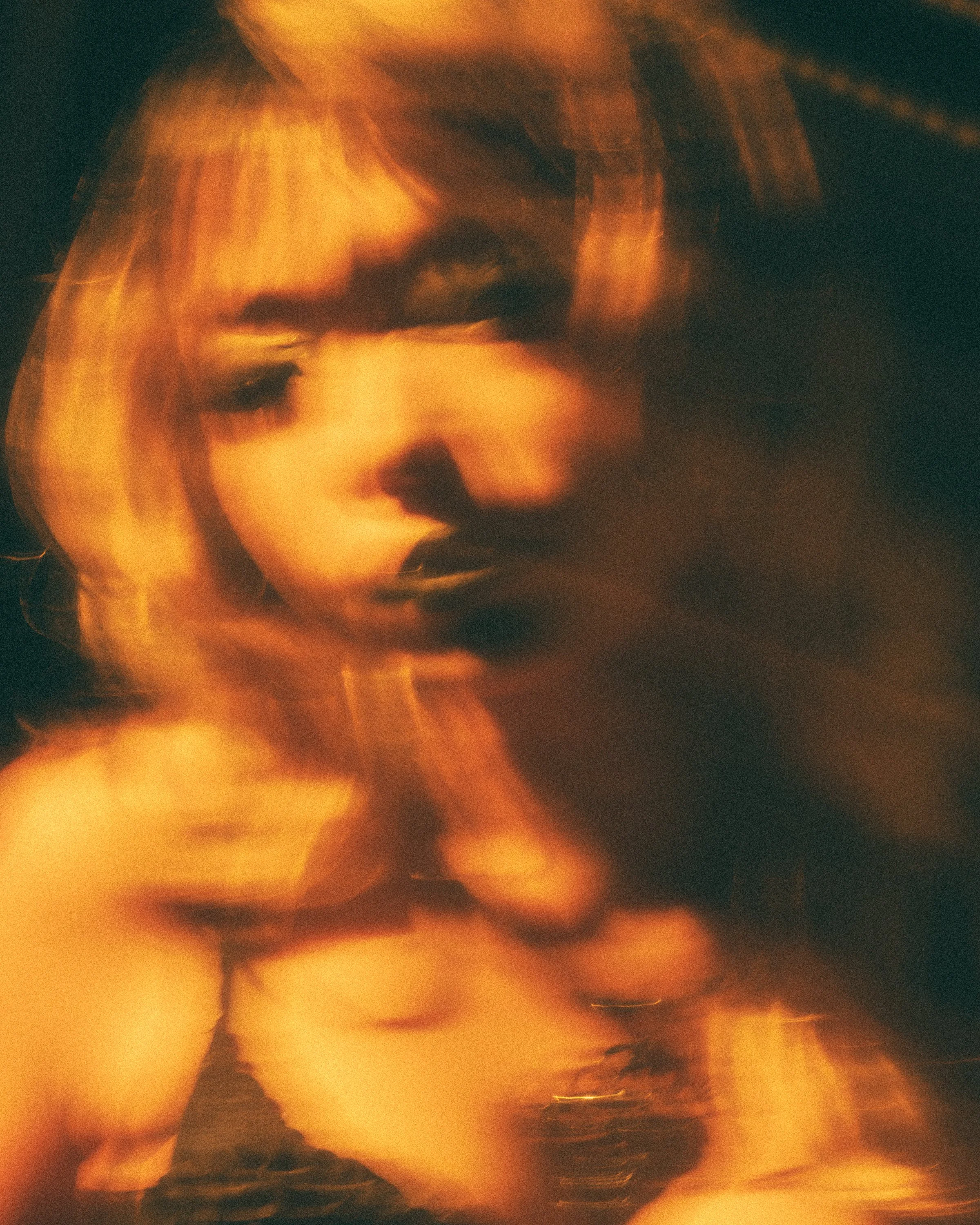 A blurry, abstract photo of a person's face illuminated by warm, orange light, with indistinct features and dark shadows.