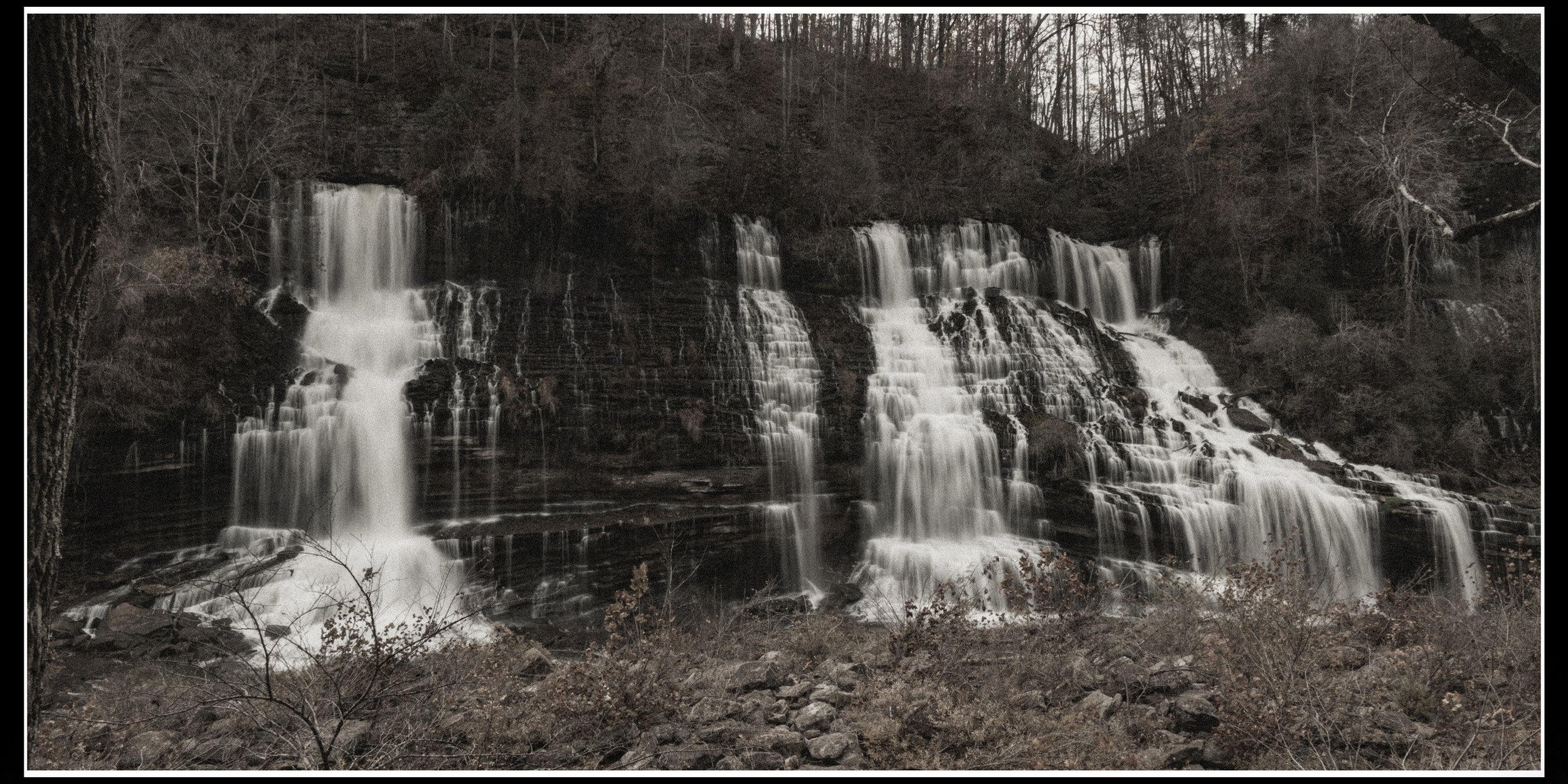A waterfall cascading over multiple rocky ledges in a forested area during winter, with leafless trees surrounding it.