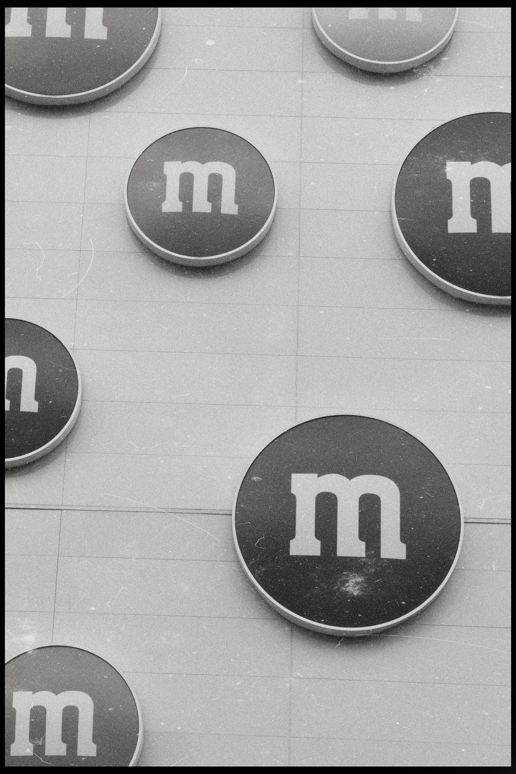Black and white photograph of several round M&M's containers with the blue M&M logo, arranged on a tiled surface.