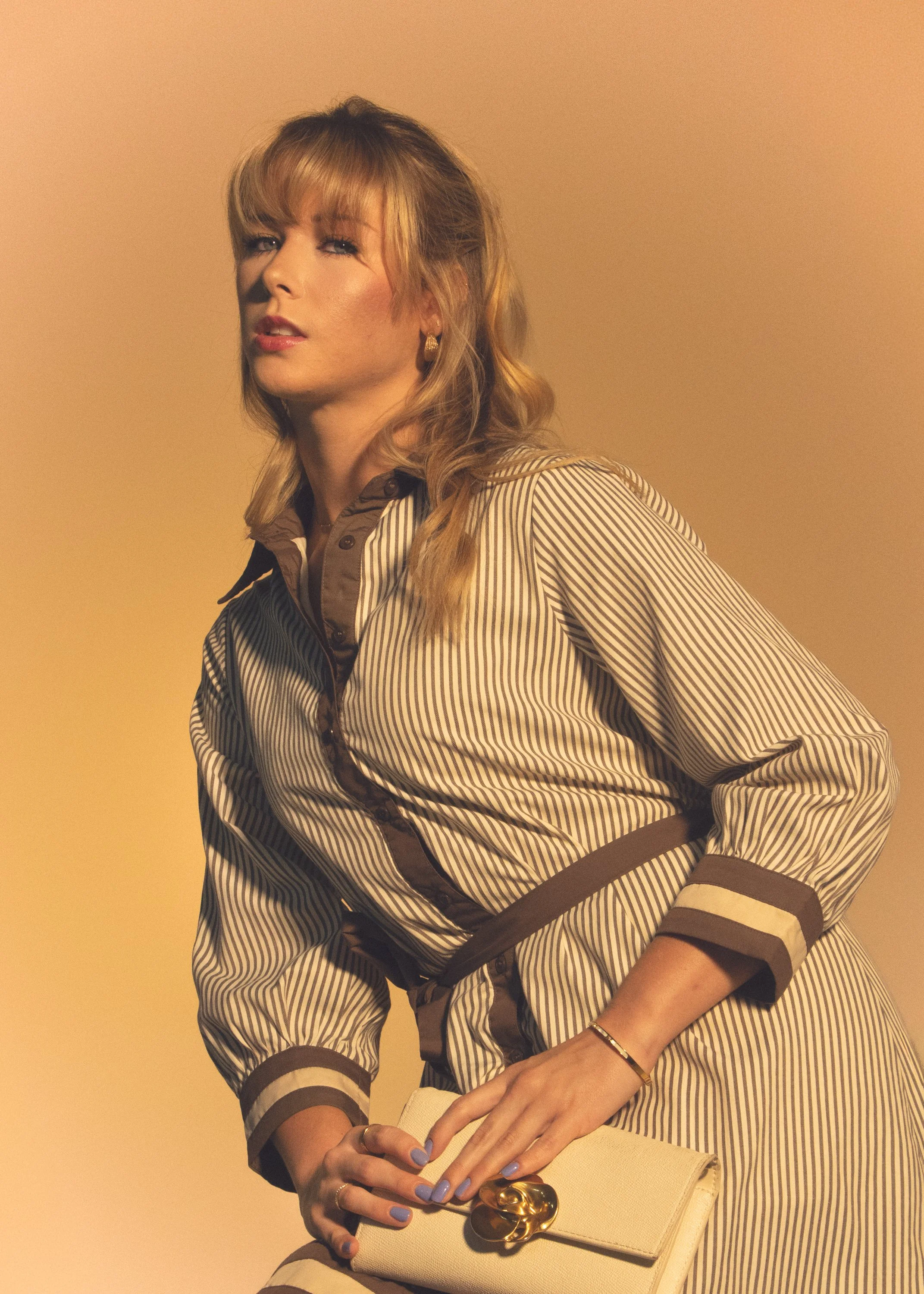Woman with blonde hair wearing a striped beige and brown outfit, holding a beige clutch with a gold ornament, standing against a yellow background.