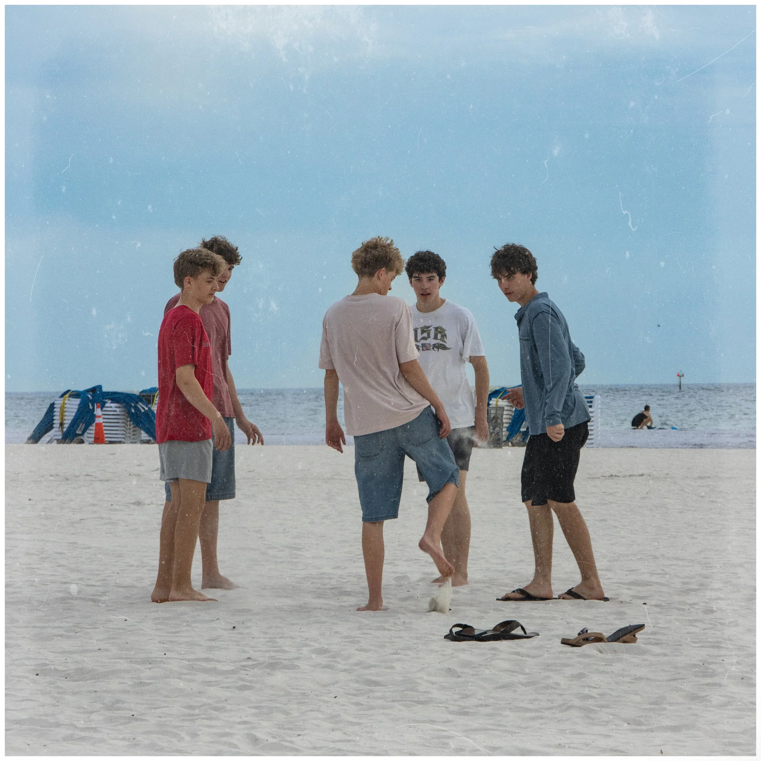 Five young men standing barefoot on a sandy beach engaged in a conversation. They are dressed casually in t-shirts and shorts. In the background, there are lounge chairs and other beachgoers, with the ocean and a cloudy sky above.