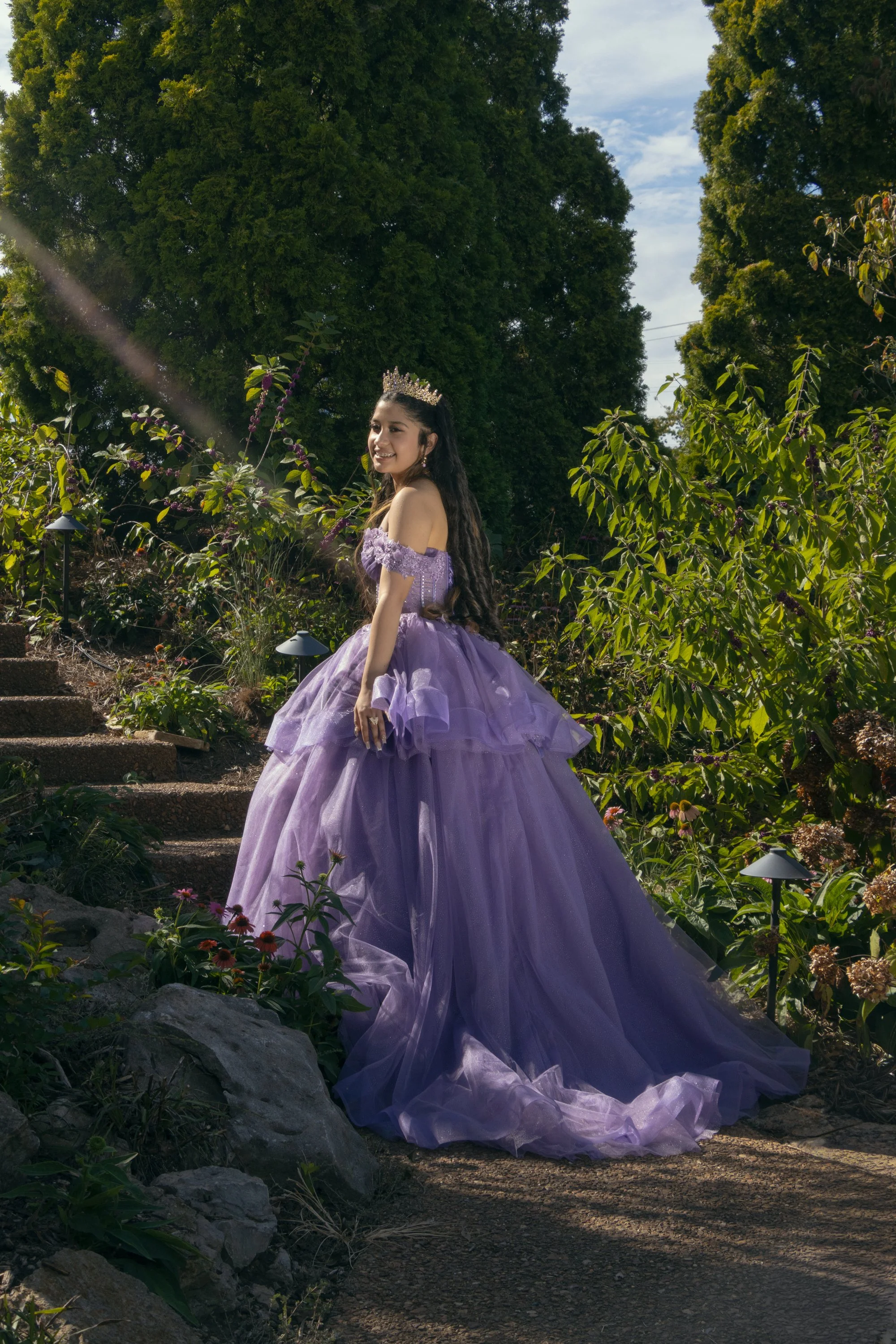Young woman in a purple gown and crown standing on garden steps with greenery and blue sky in background.