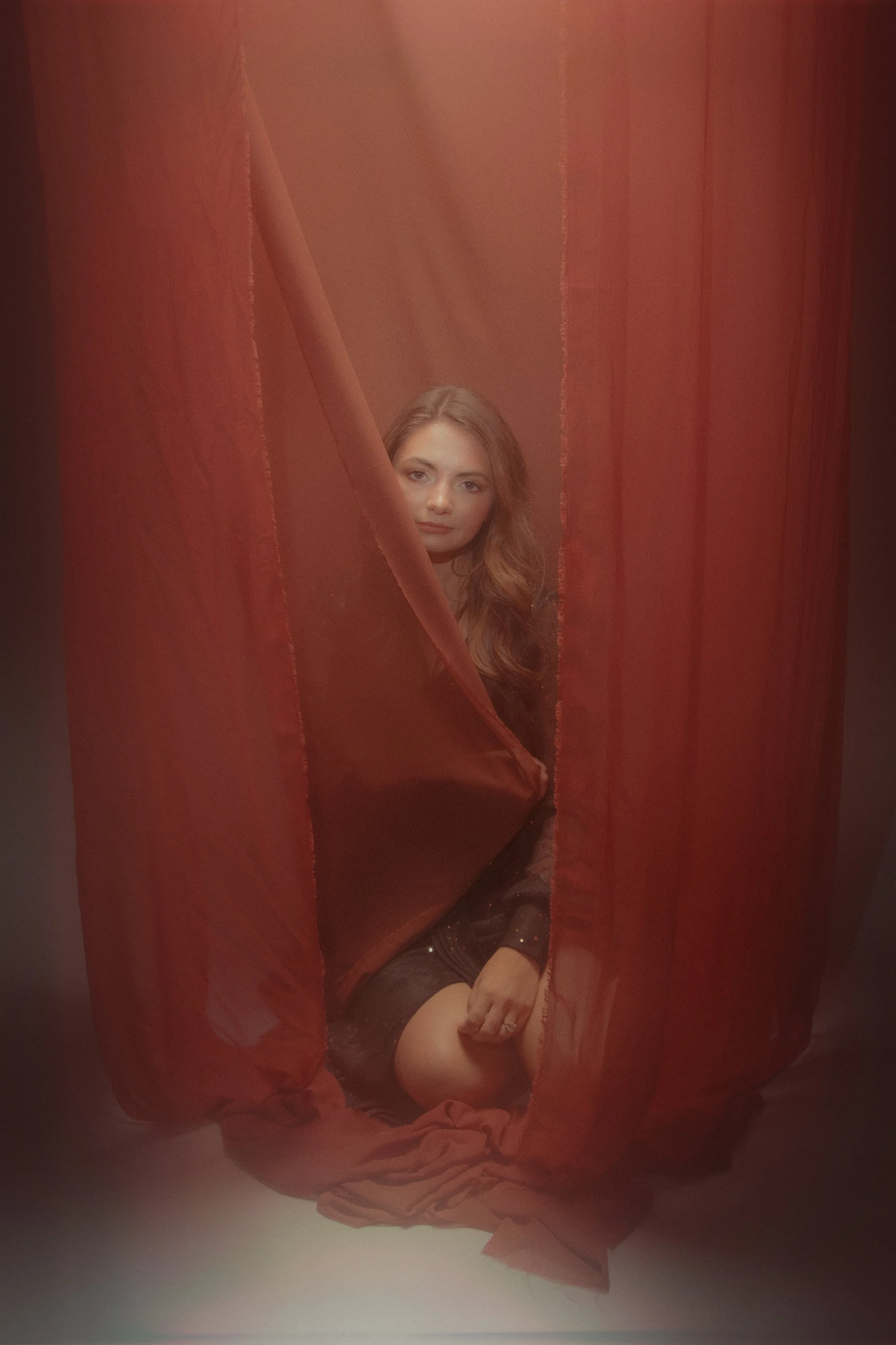 A woman with long wavy hair sitting behind a red curtain with a soft expression on her face.