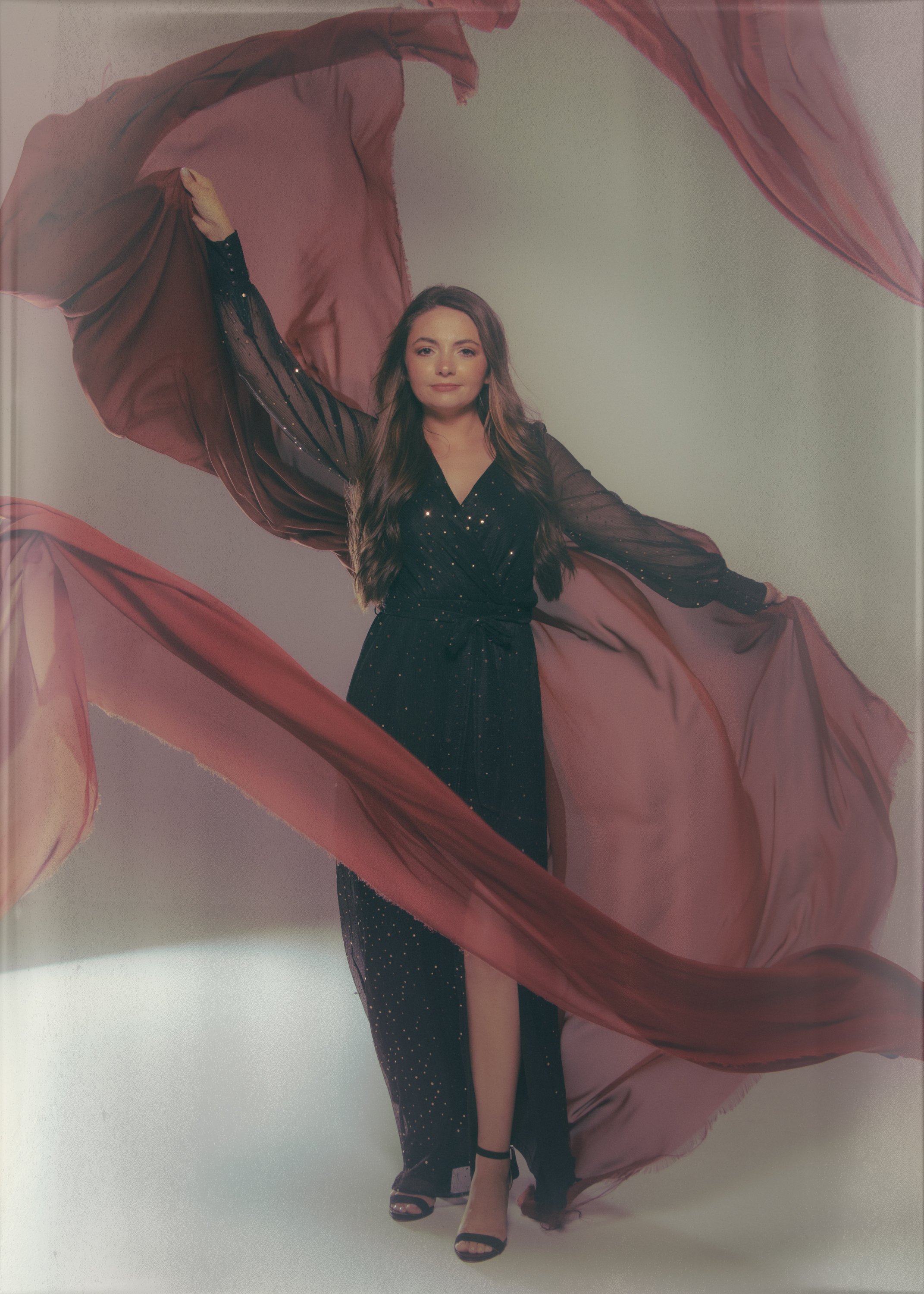 A woman with long, wavy brown hair wearing a black dress with sheer sleeves and small rhinestones, standing against a plain white background, holding pink fabric that flows around her and partially covers her legs. She is wearing black heeled sandals