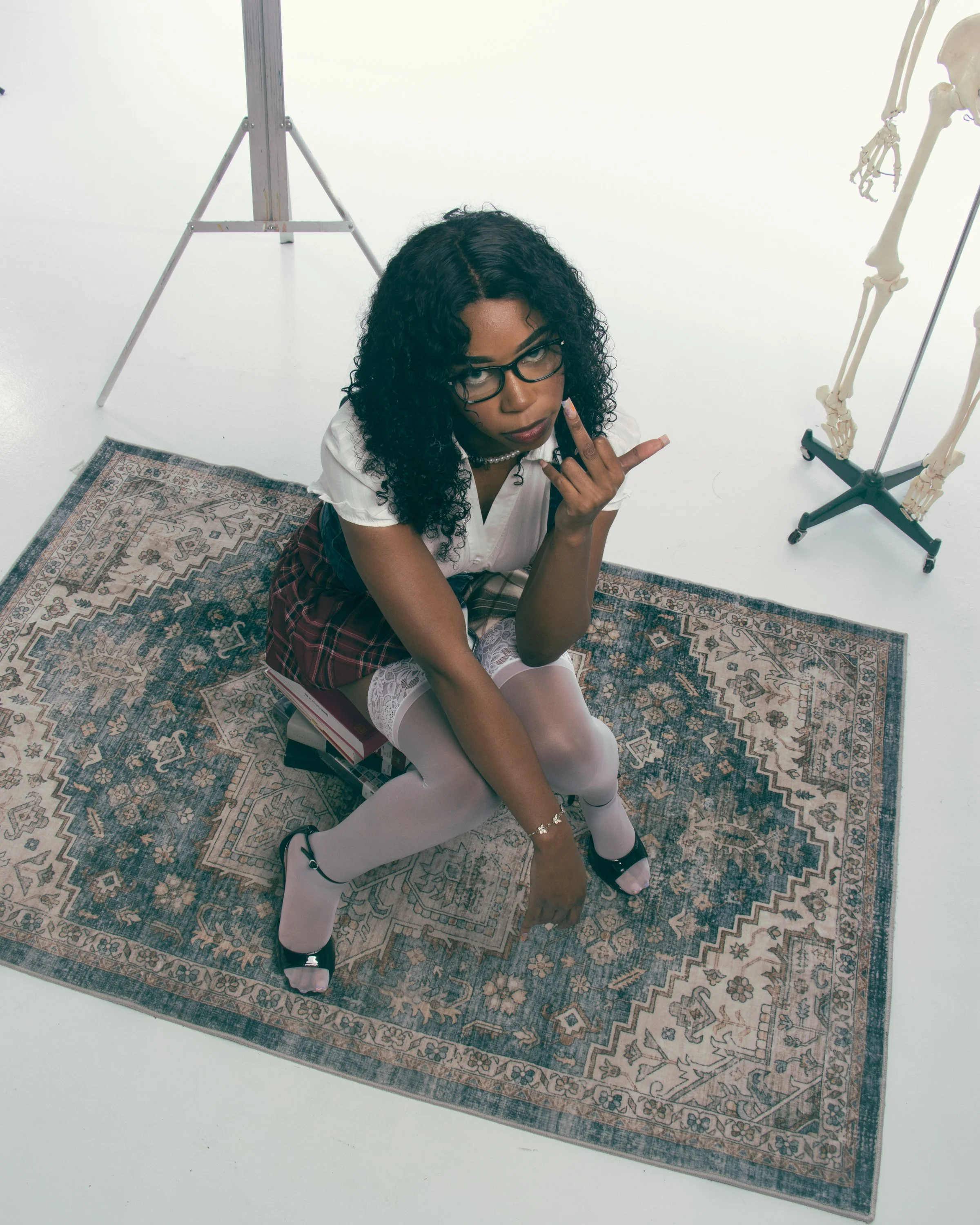 A woman with curly black hair and glasses sitting on a rug on the floor on her knees, making a hand gesture with her middle finger and thumb extended. She is dressed in a white top, plaid skirt, white tights, and black shoes with white socks. Behind 