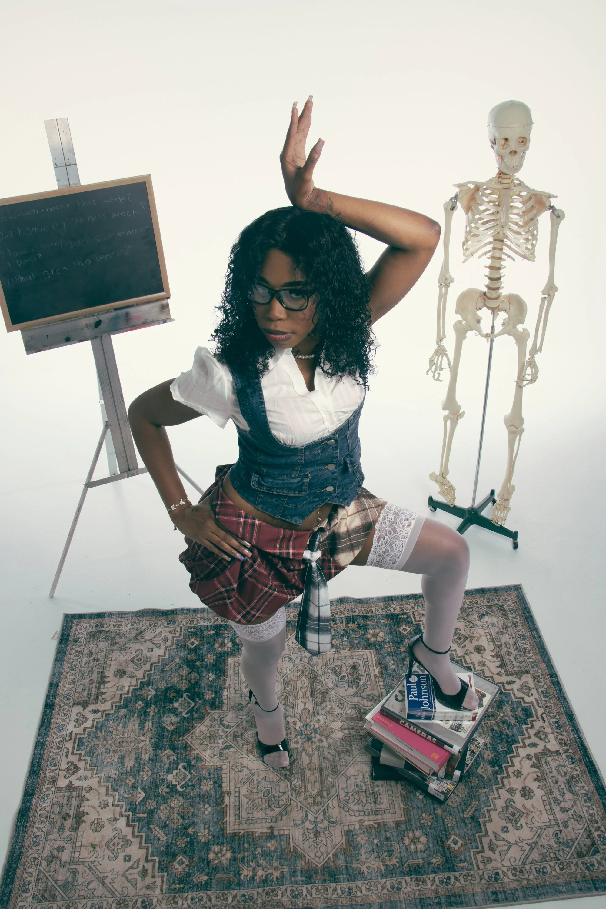 A woman dressed in a schoolgirl costume with white stockings, a plaid skirt, and a white blouse, is striking a dramatic pose on a vintage rug, standing on a stack of photography and camera guide books. She is surrounded by a skeleton model and an eas