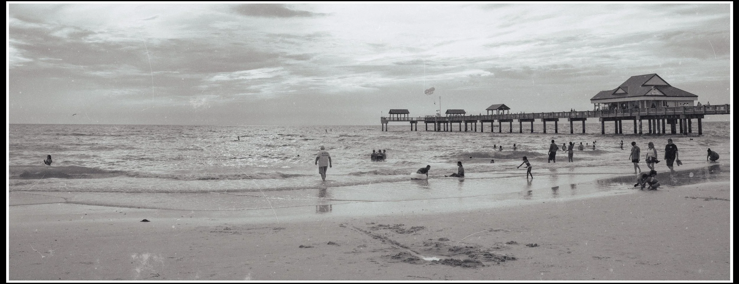 People enjoying a day at the beach with a pier in the background, some swimming, sitting, and walking along the shore, under a cloudy sky.