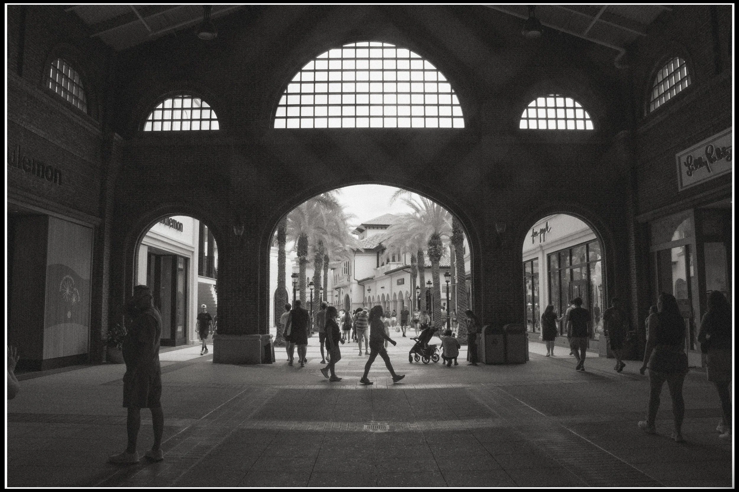 People walking through an outdoor shopping area with palm trees, storefronts, and a covered archway with large windows.