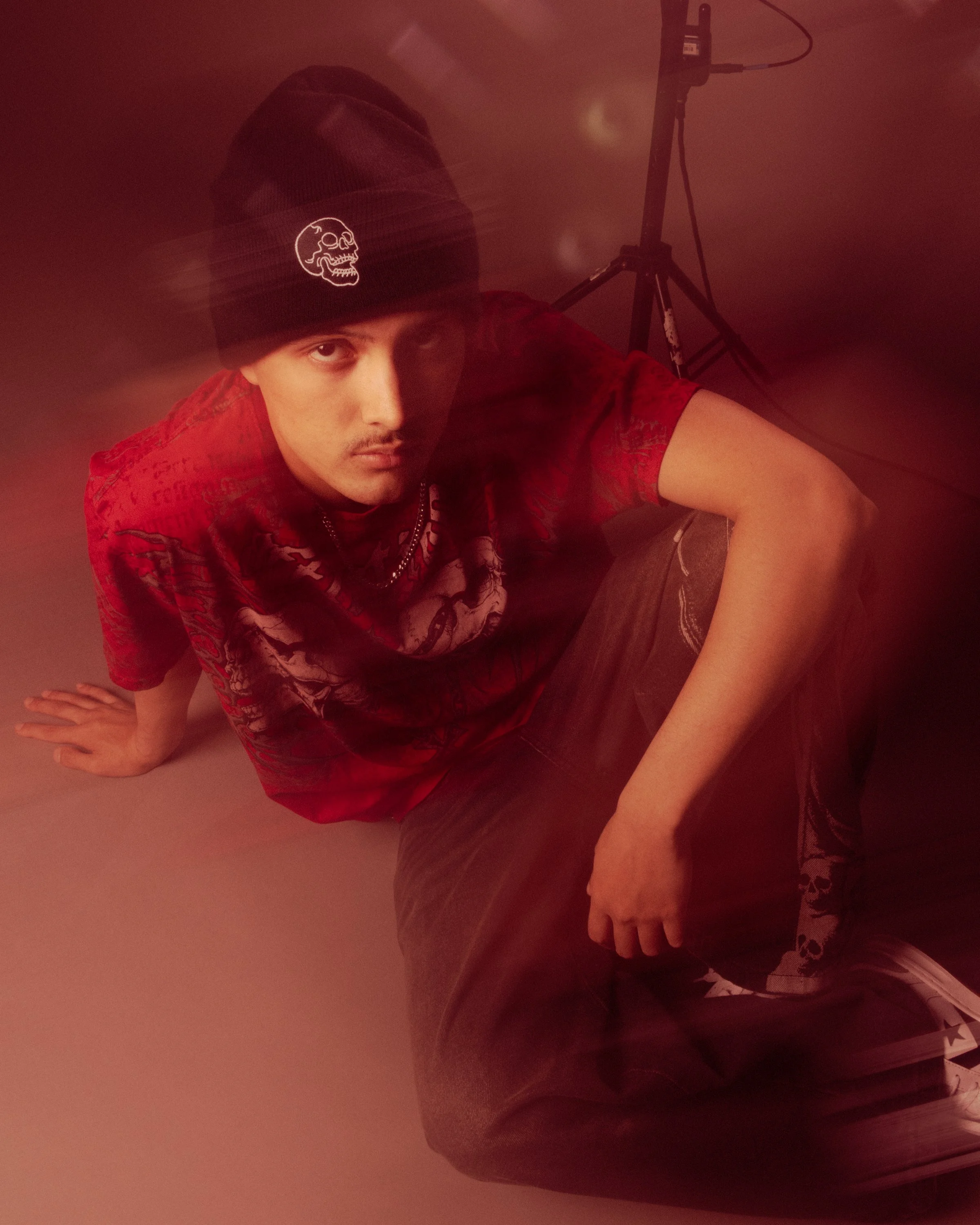 A person taking a selfie, wearing a black beanie with a skull logo, a red shirt, gray shorts, and sitting on the floor with a red ambient light. There is a tripod in the background.