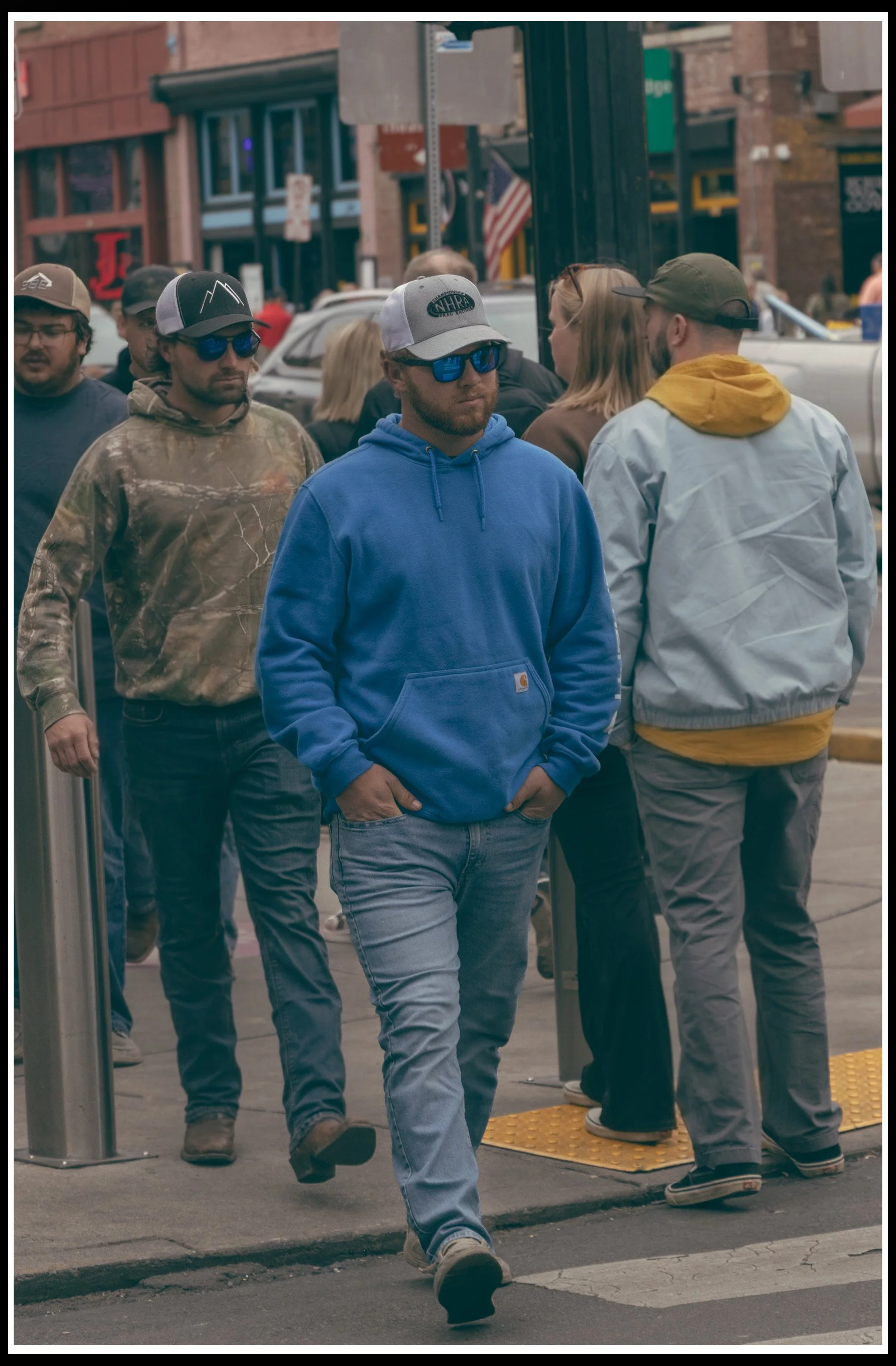 People waiting in line outside a store in an urban area, some wearing sunglasses and casual clothing, with storefronts and parked cars visible in the background.