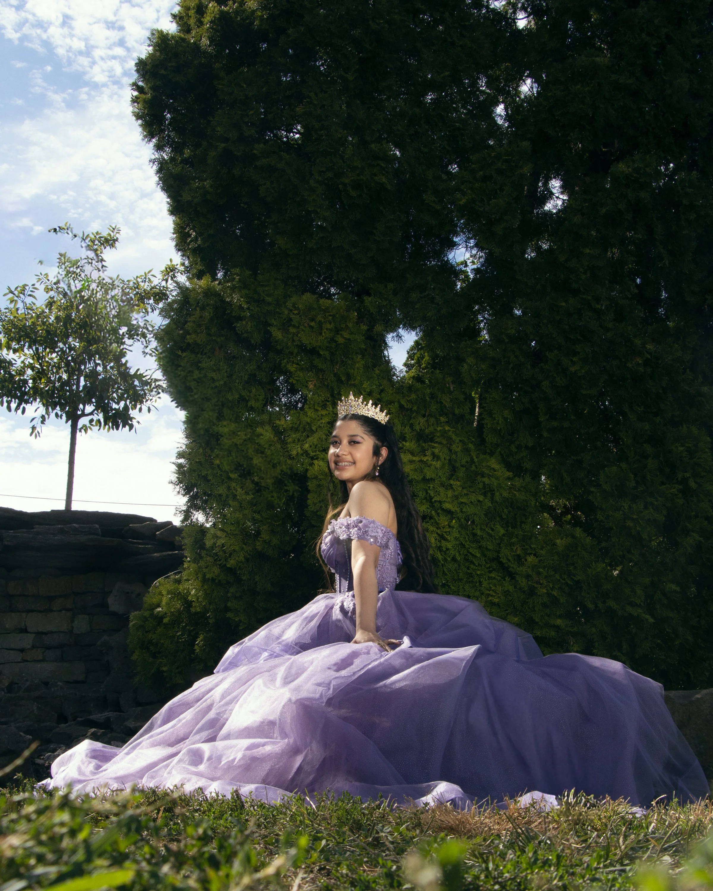 A young woman dressed in a purple gown and wearing a tiara is kneeling outdoors on grass with a large tree behind her. The sky is partly cloudy.