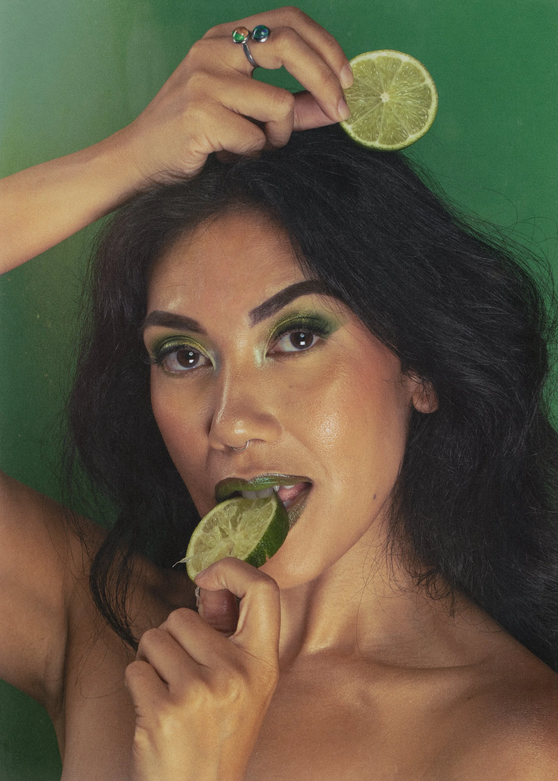 A woman with black hair and vibrant green eye makeup holding a lime slice in her mouth, another lime slice on her forehead, and gripping a lime wedge with her fingers. She is against a green background.