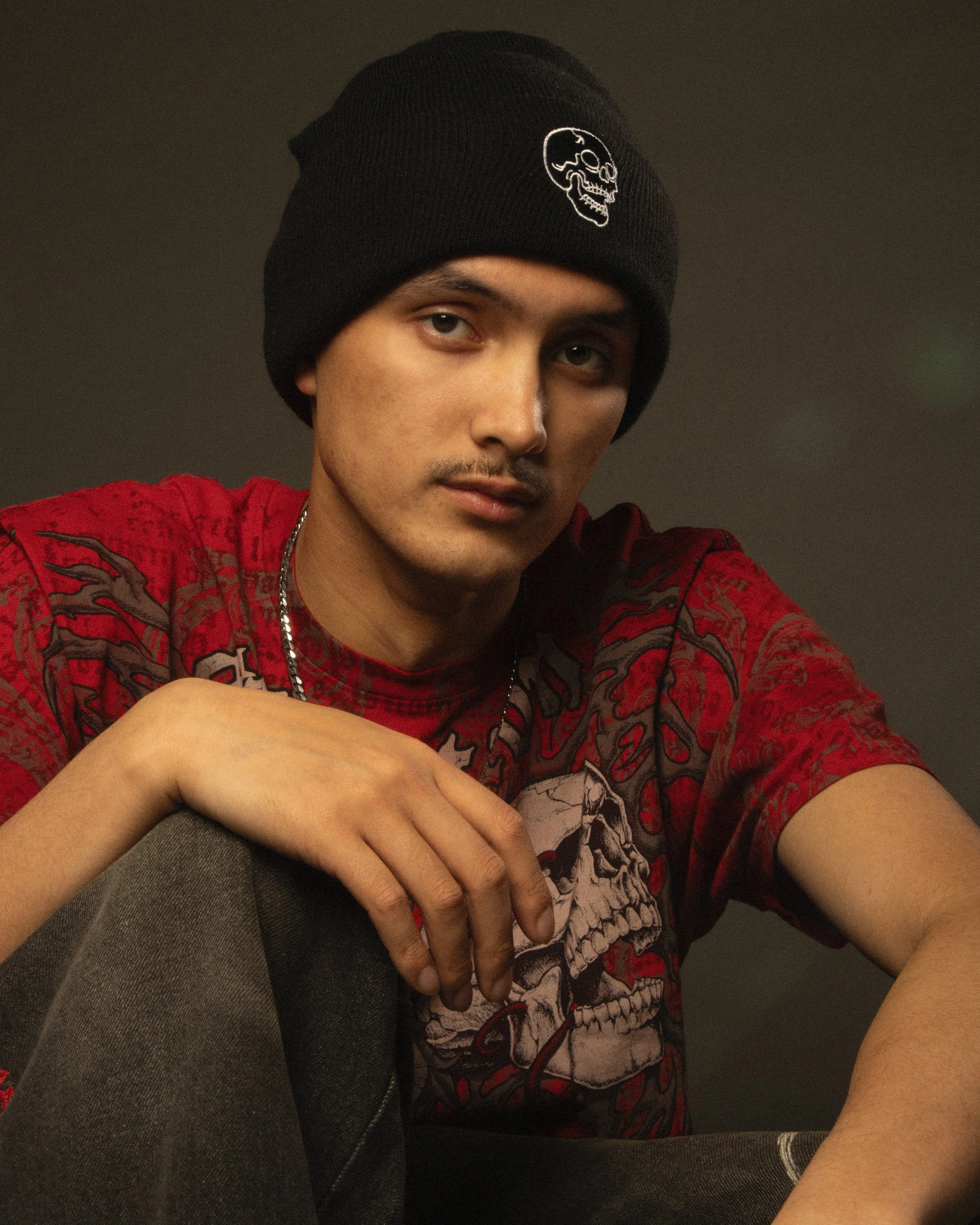 A young man wearing a black beanie with a skull print, a red graphic T-shirt, and a silver chain, seated with his arm resting on his knee, in a dimly lit room.