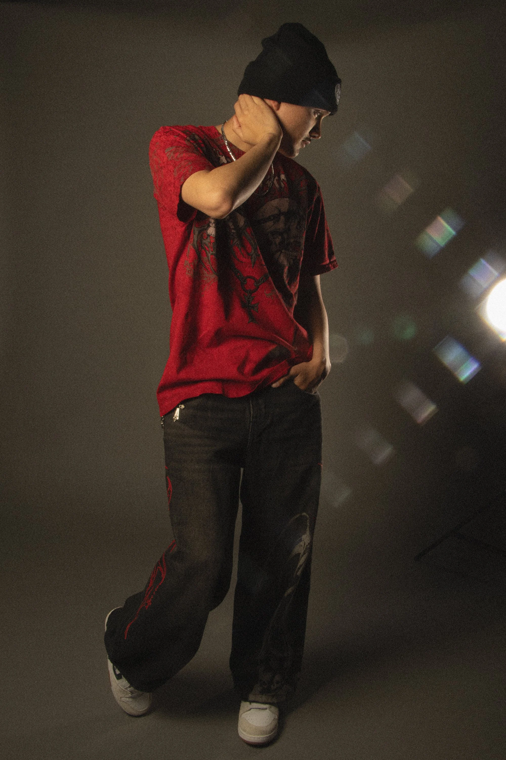 A person wearing a black beanie, red and black graphic shirt, black ripped jeans with red stripe detail, and white sneakers, standing against a dark background with a light source to the right.